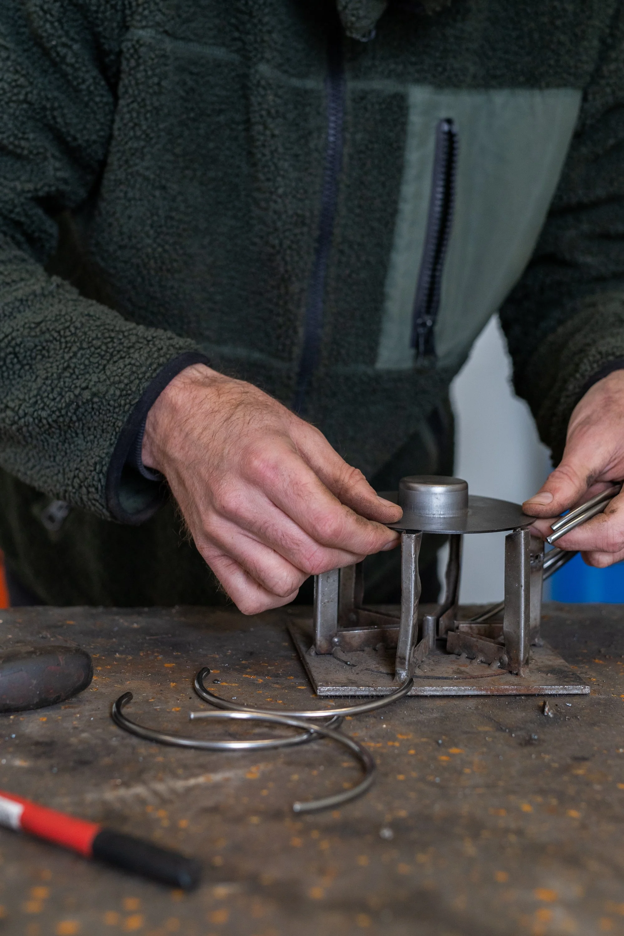 A person working on a metal project, holding a clamp and working on a circular metal component on a workbench.