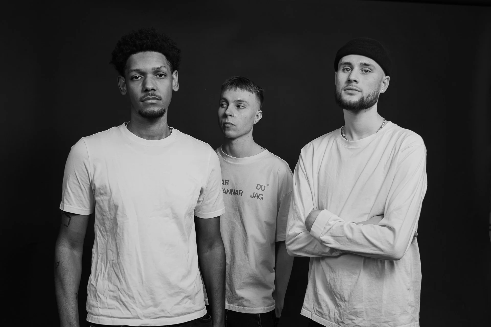 Black and white photo of three young men standing against a dark background, facing the camera with serious expressions. The man on the left has an afro hairstyle, mustache, and beard, wearing a plain t-shirt. The man in the middle has short, straigh