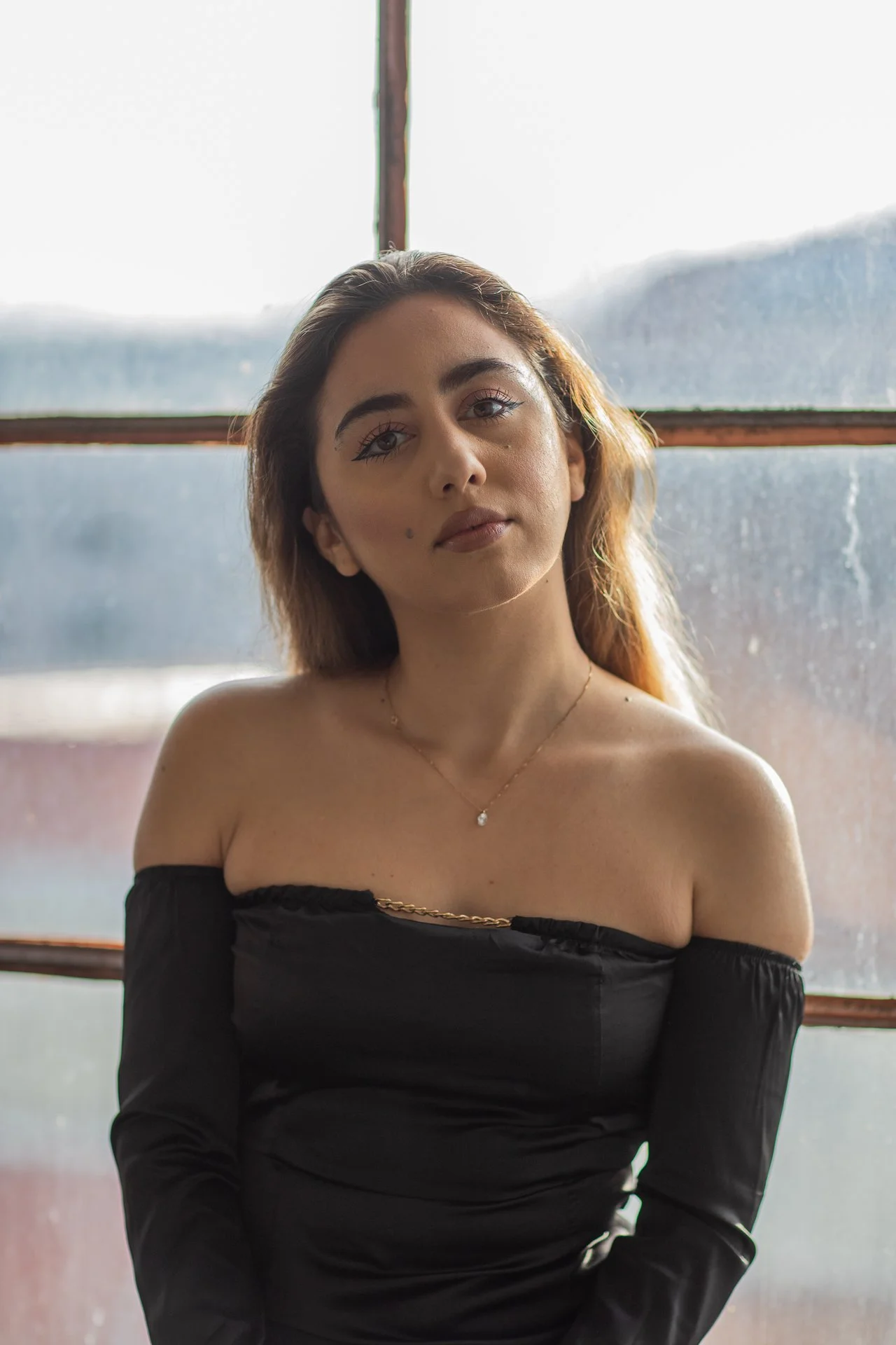 A young woman with light brown hair and makeup, wearing an off-shoulder black dress and a delicate necklace, sitting in front of a window with a blurred cityscape background.