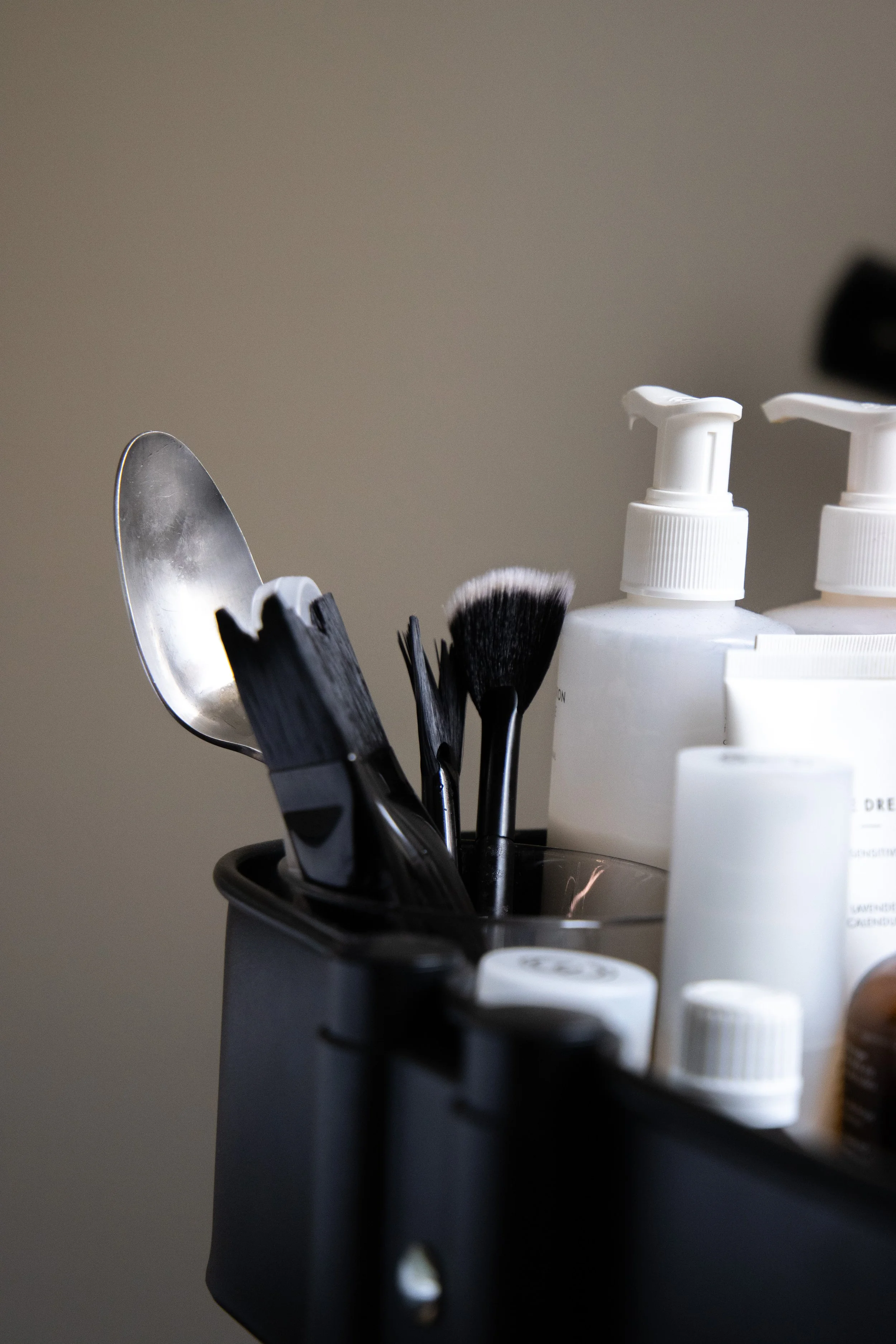 Makeup brushes, a spoon, and skincare bottles with pumps in a black container