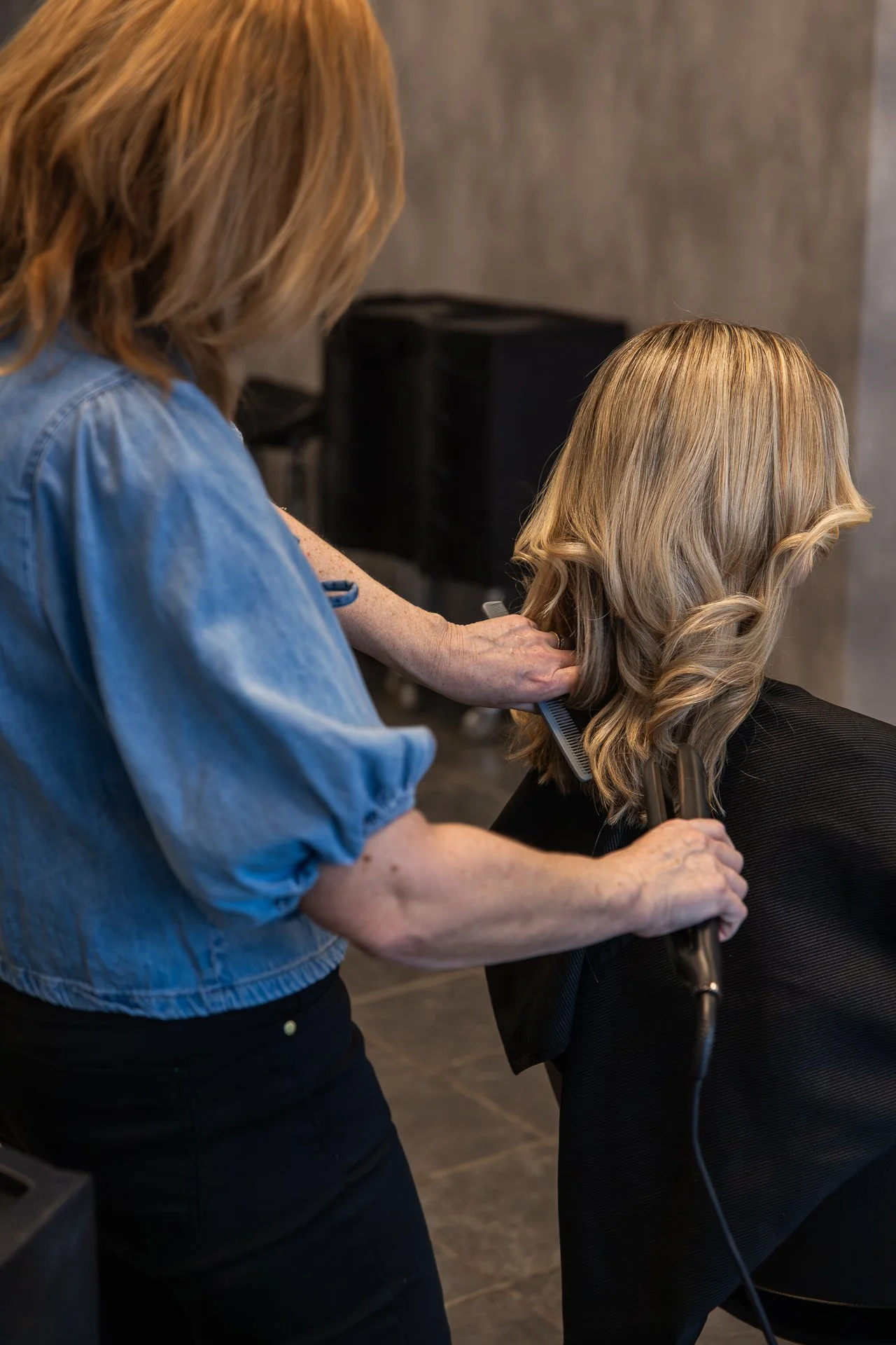 A hairstylist cuts and styles a woman's blonde hair with curling styling tools in a hair salon.
