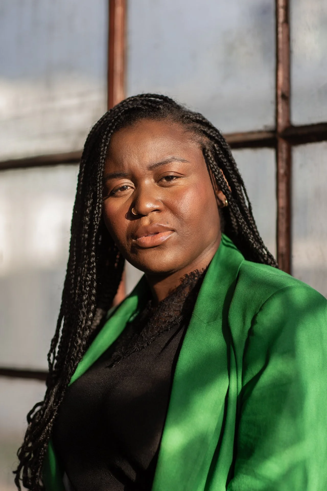 A woman with dark braided hair, wearing a black top and a bright green blazer, standing in front of a metallic background.