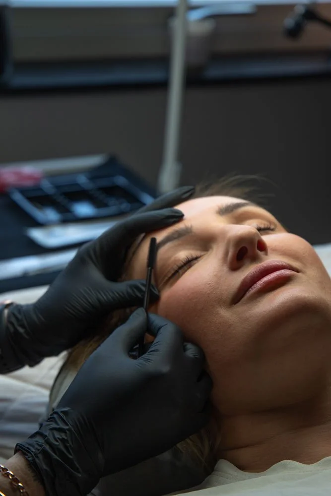 A woman receives a cosmetic eyebrow tattooing or microblading procedure while lying down, with a technician wearing black gloves using a small tool on her eyebrow.