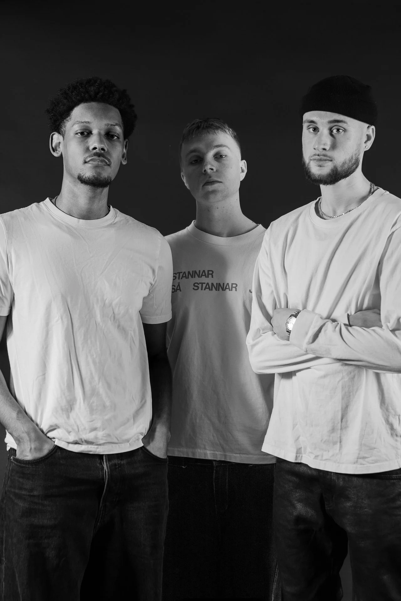 Black and white photo of three young men standing together against a dark background, wearing casual white t-shirts and looking at the camera with serious expressions.