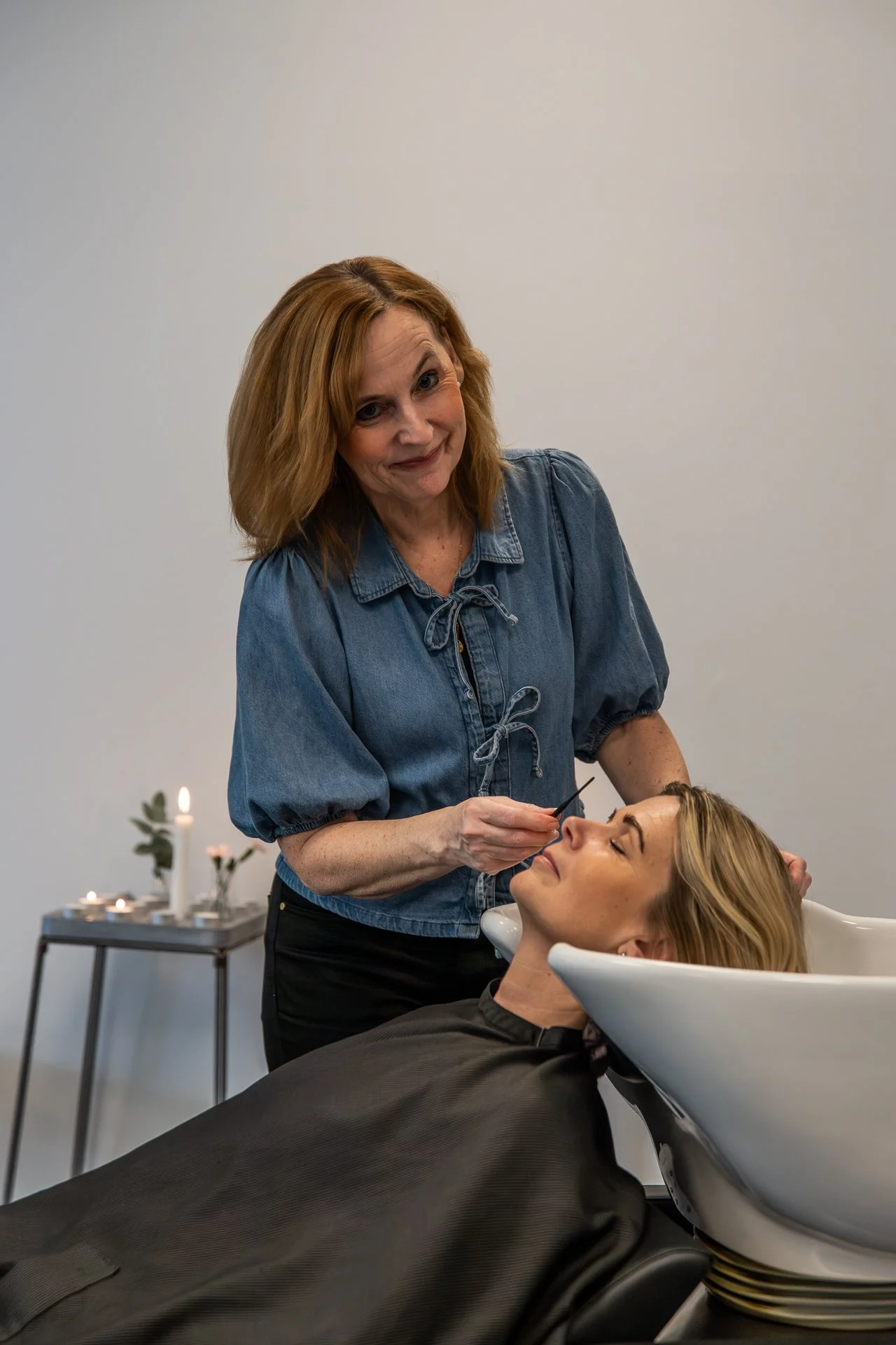 A woman lying back in a salon sink with her head tilted, receiving a beauty treatment, while a stylist applies makeup or cosmetic product to her face. The room has a white wall and a table with candles and flowers in the background.