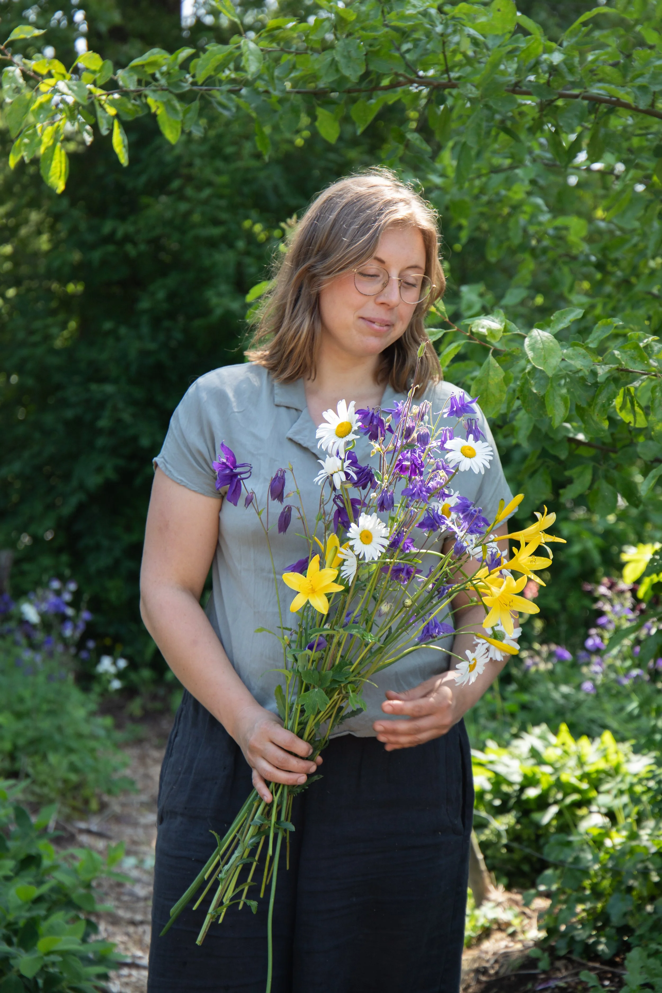 A woman standing outdoors in a garden holding a large bouquet of mixed flowers, including daisies, yellow lilies, and purple flowers, with green foliage in the background.