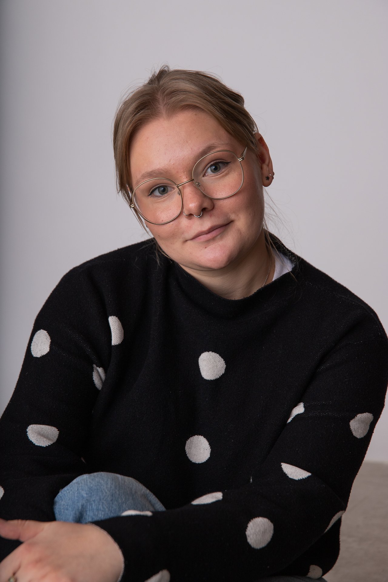 A young woman with short reddish hair, glasses, and a septum piercing wearing a black sweater with white polka dots, sitting against a plain light gray background.