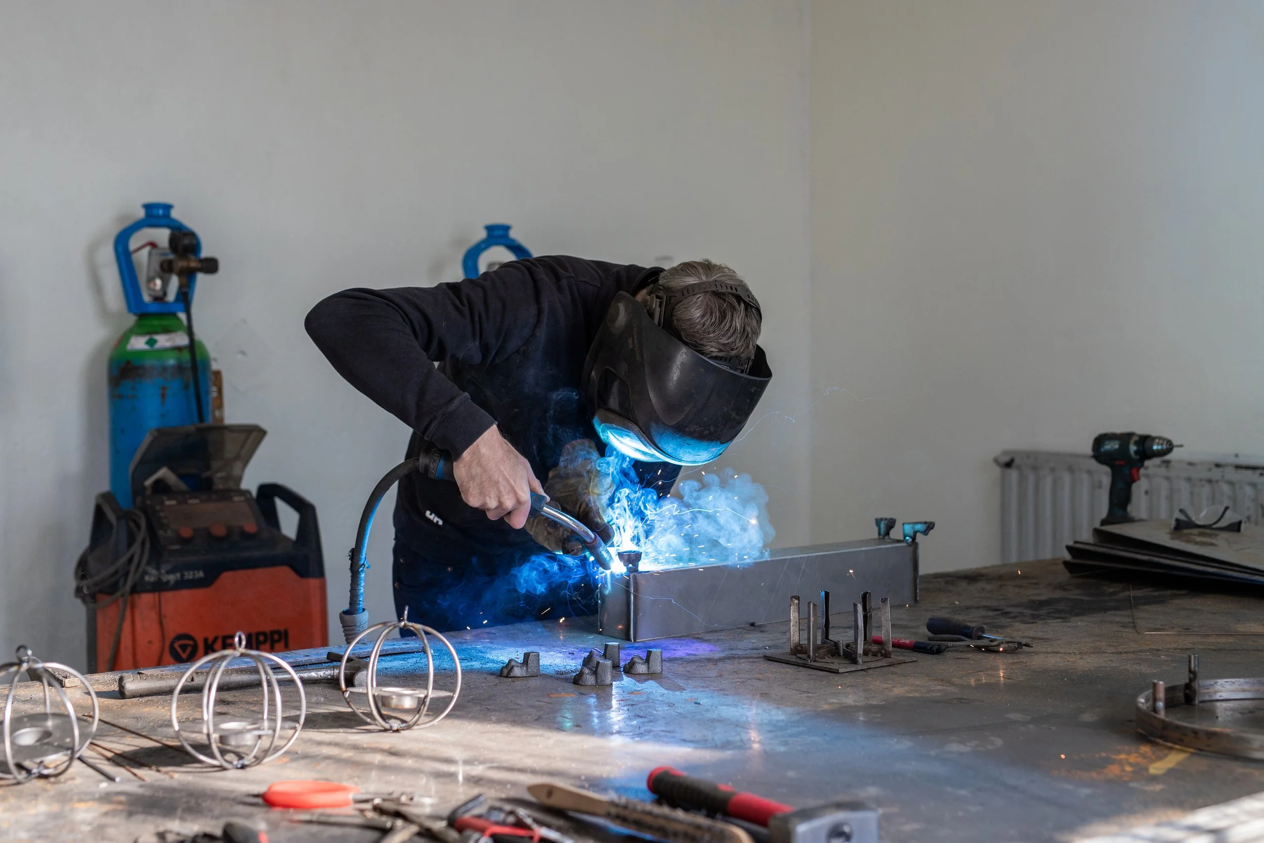A person welding metal on a workbench, wearing a protective helmet and gloves, with sparks and smoke from the welding process.
