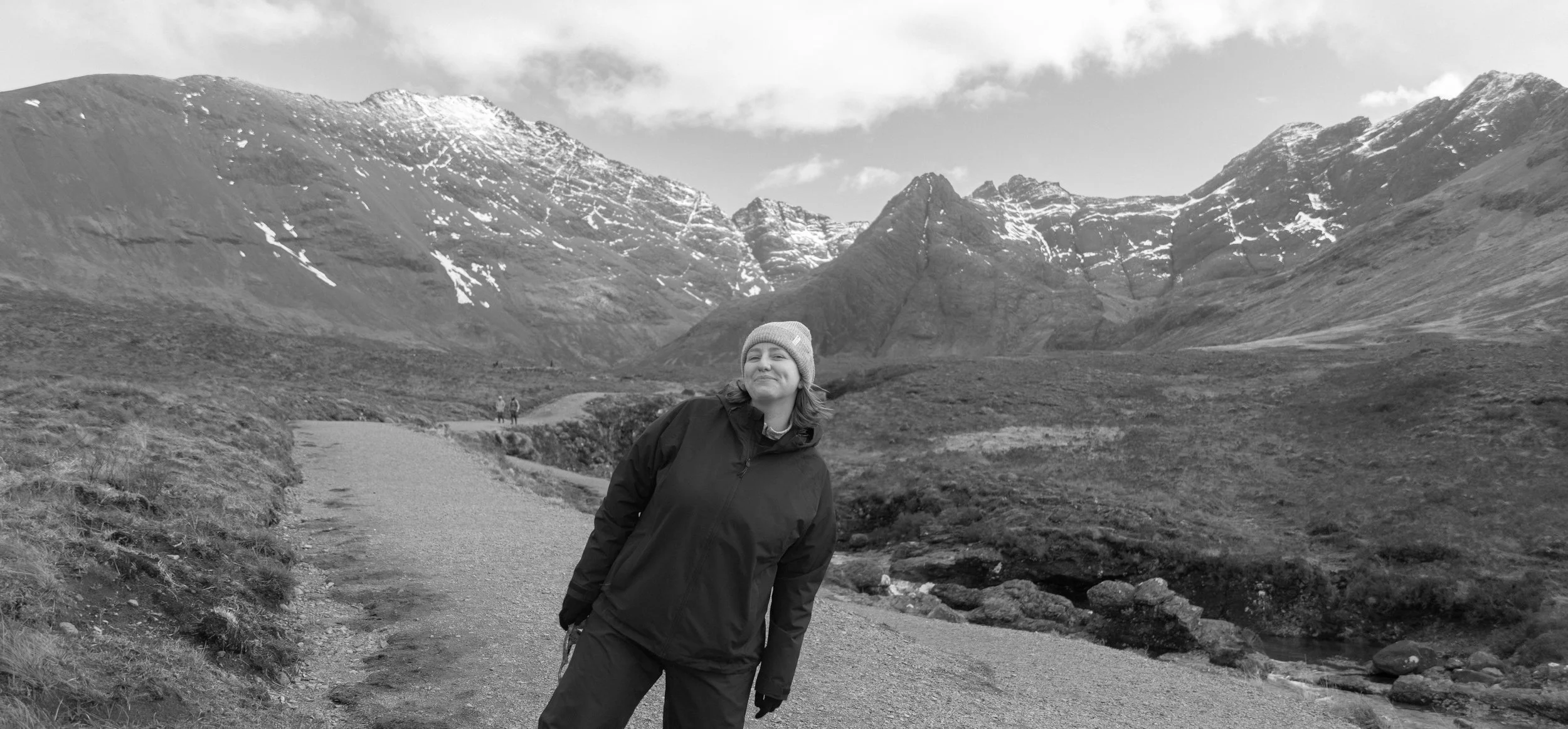 A woman standing on a mountain trail in a mountainous landscape with snow-capped peaks in the background.