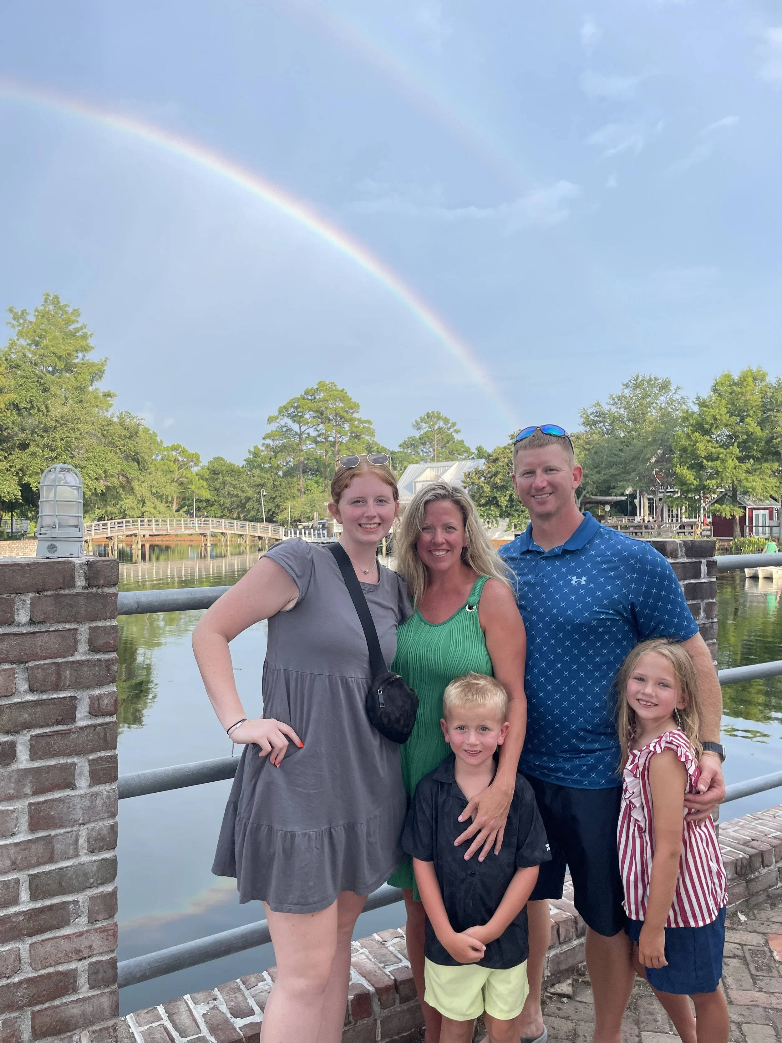 A family of six posing by a canal with a rainbow in the sky, surrounded by green trees and a small bridge in the background.