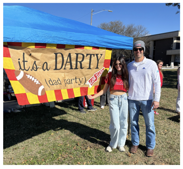 A man and woman standing outside next to a colorful banner that reads 'it's a DARTY (dad party) for dad'. They are smiling, wearing casual clothing, and posing for the photo on a sunny day.