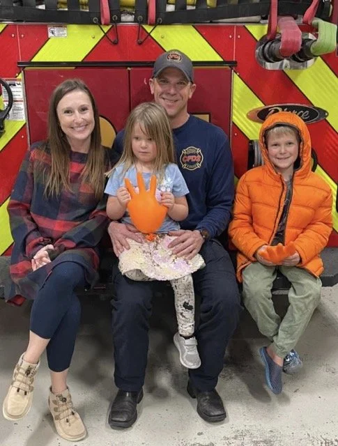 Family of four sitting inside a fire truck with a red and yellow background. The mother has long brown hair and a plaid shirt, the father is in a navy blue shirt and cap, the girl in the middle has long hair, a blue shirt, and is holding an orange glove, and the boy on the right is in an orange jacket and holding gloves.