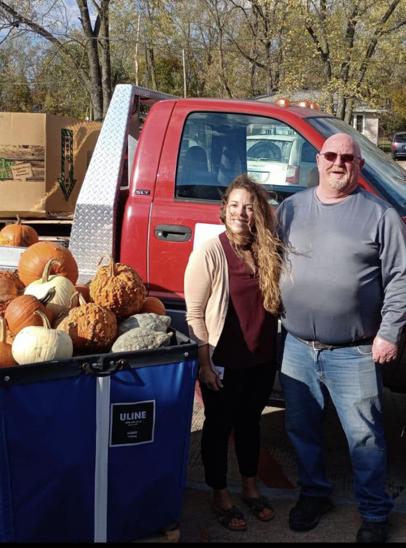 Two people standing next to a truck with a bin of pumpkins at an outdoor pumpkin patch.
