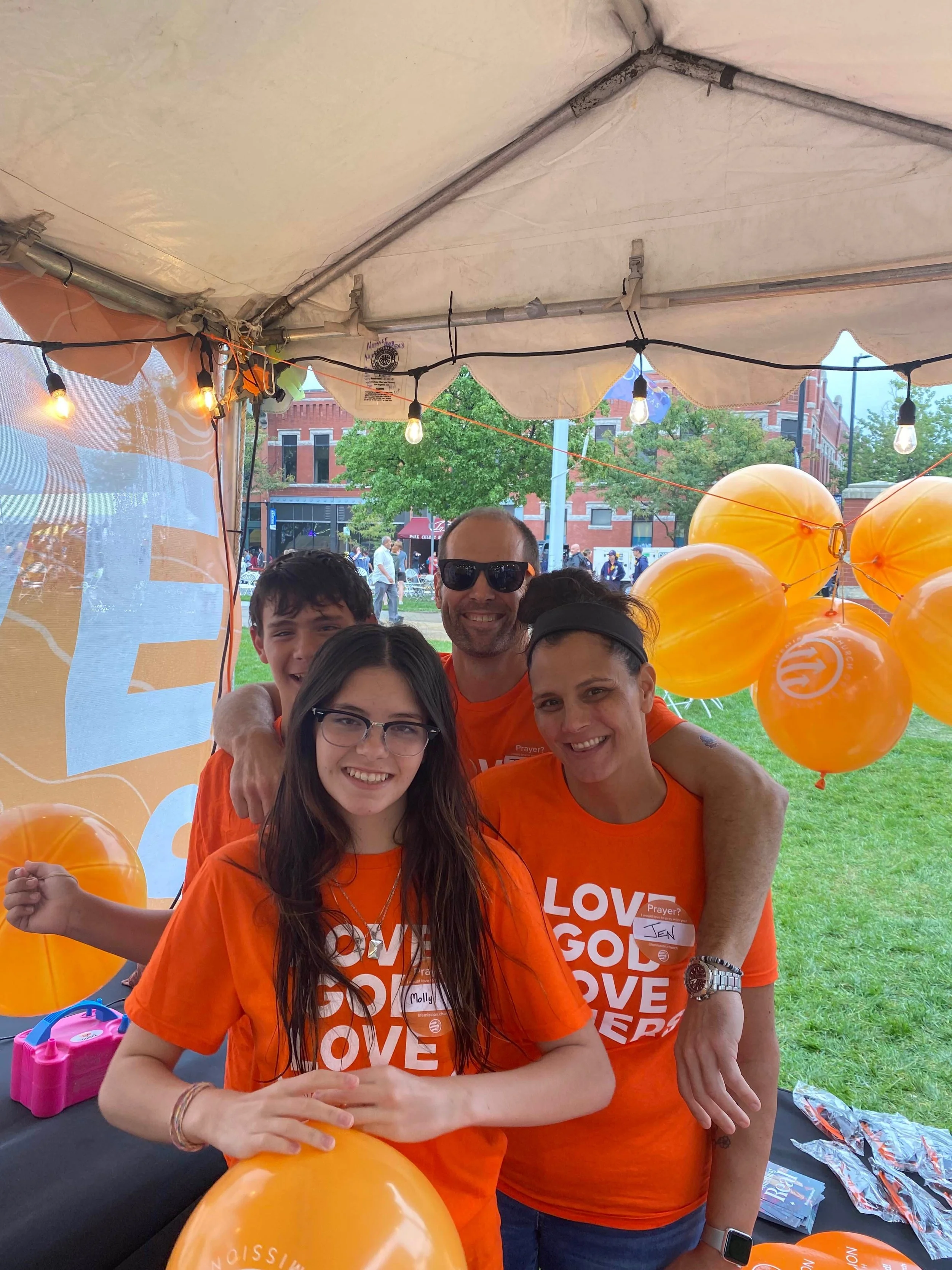 People at an outdoor event wearing matching orange shirts with the words 'LOVE GOD LOVE OTHERS,' holding orange balloons, under a white canopy, with a city street and red-brick buildings in the background.