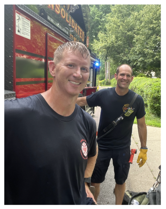 Two firefighters stand outside a red fire truck with equipment, smiling at the camera. The firefighter in the foreground has short light brown hair and wears a navy blue T-shirt. The other firefighter has a bald head, wears a navy blue T-shirt, yellow gloves, and has a radio clipped to his chest.