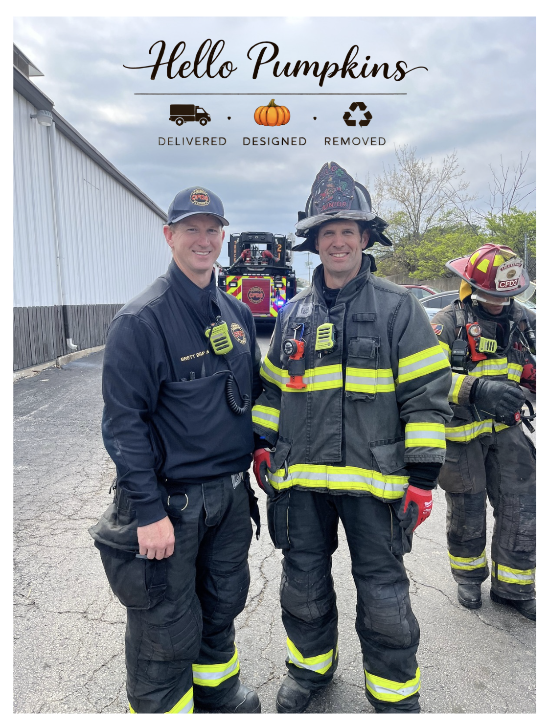 Two firefighters in uniform standing next to each other outside, with a fire truck behind them. Both are smiling. One is wearing a firefighter helmet, and the other firefighter's helmet is on his head. The sky is cloudy.