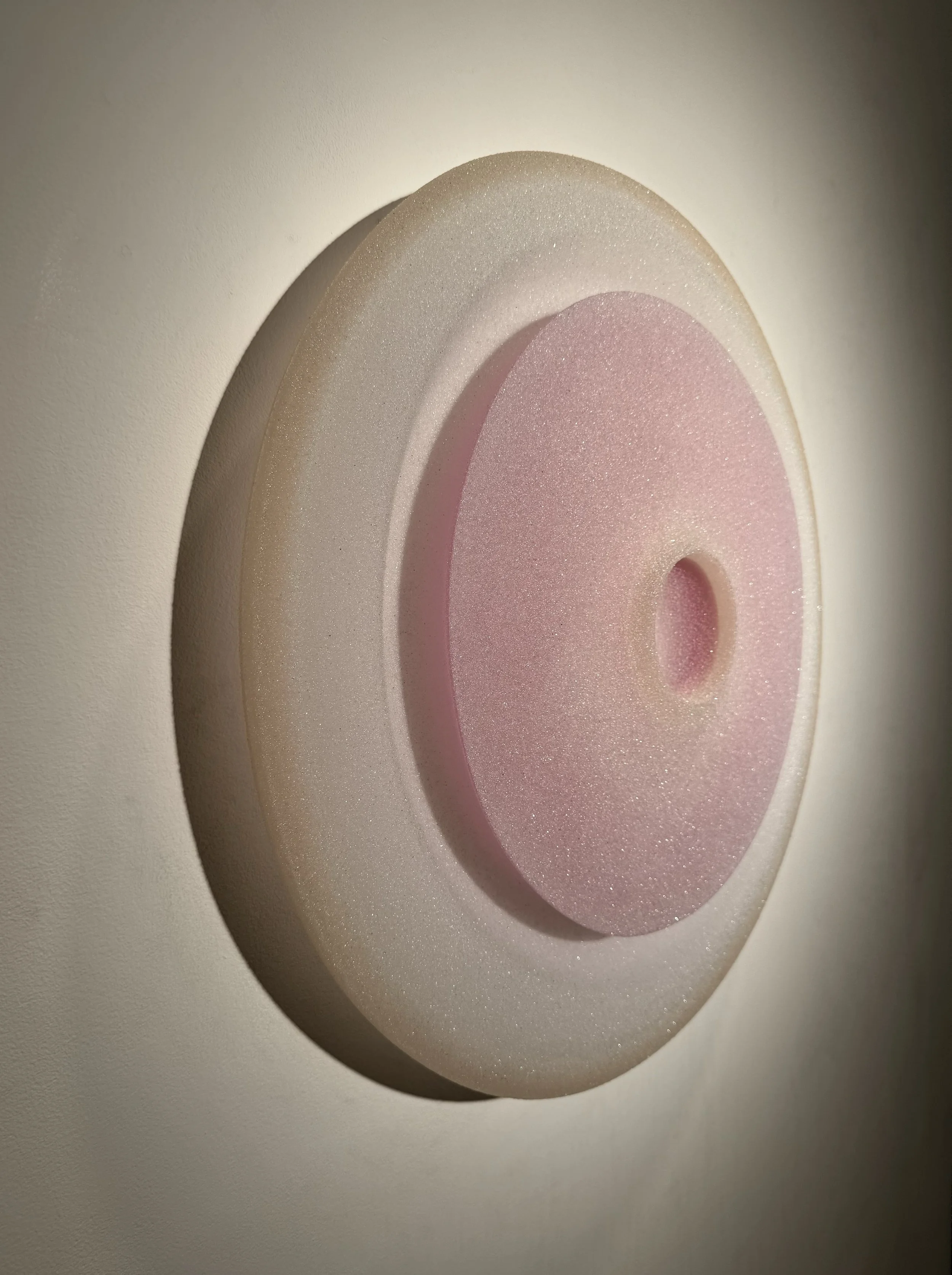 A symmetrical, circular, abstract sculpture with a pink center, beige middle layer, and white outer layer, mounted on a light wall.