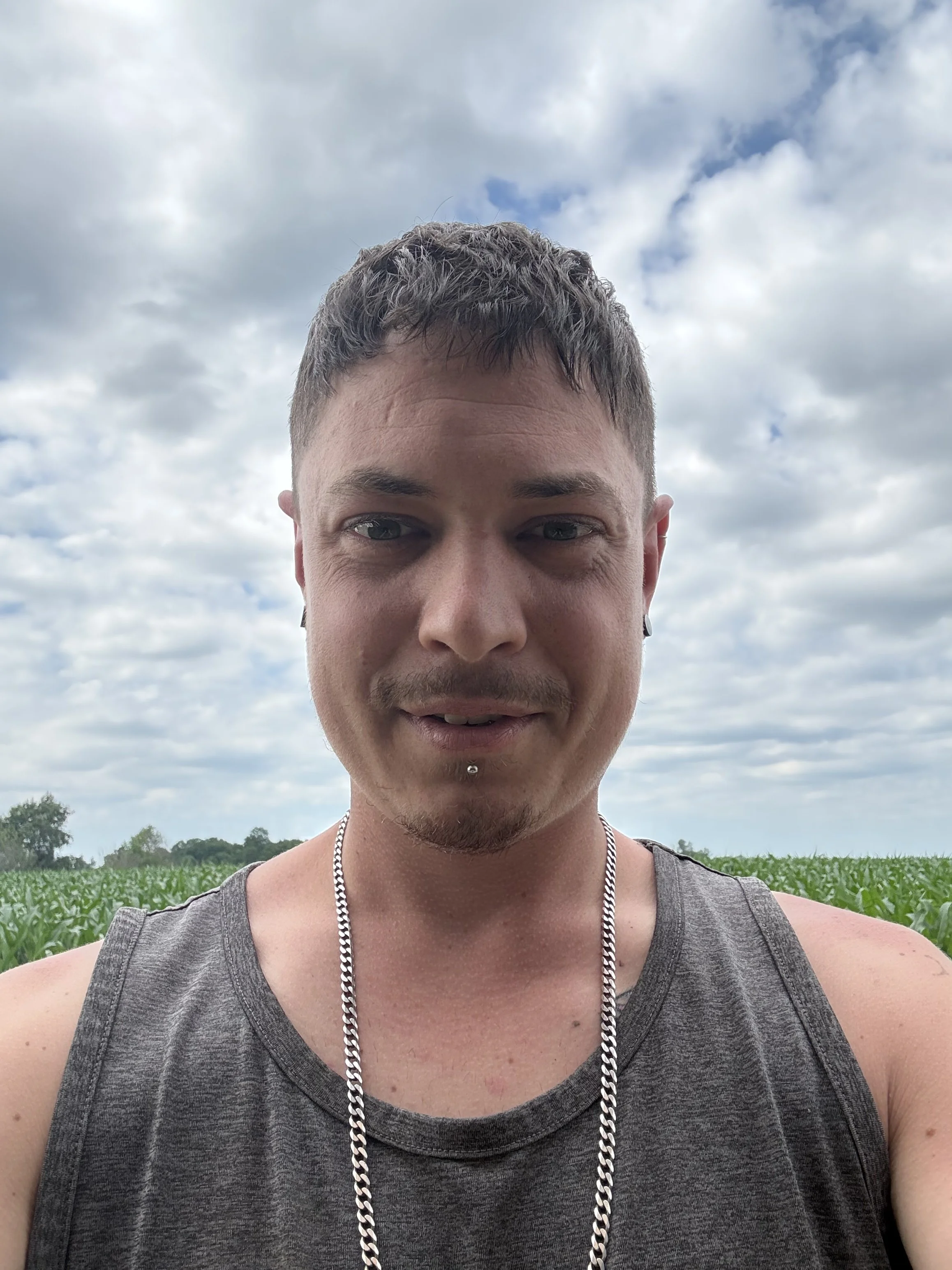 A young man with short brown hair and a piercing below his lower lip, wearing a sleeveless gray shirt and a silver chain necklace, standing outdoors against a cloudy sky with green fields in the background.