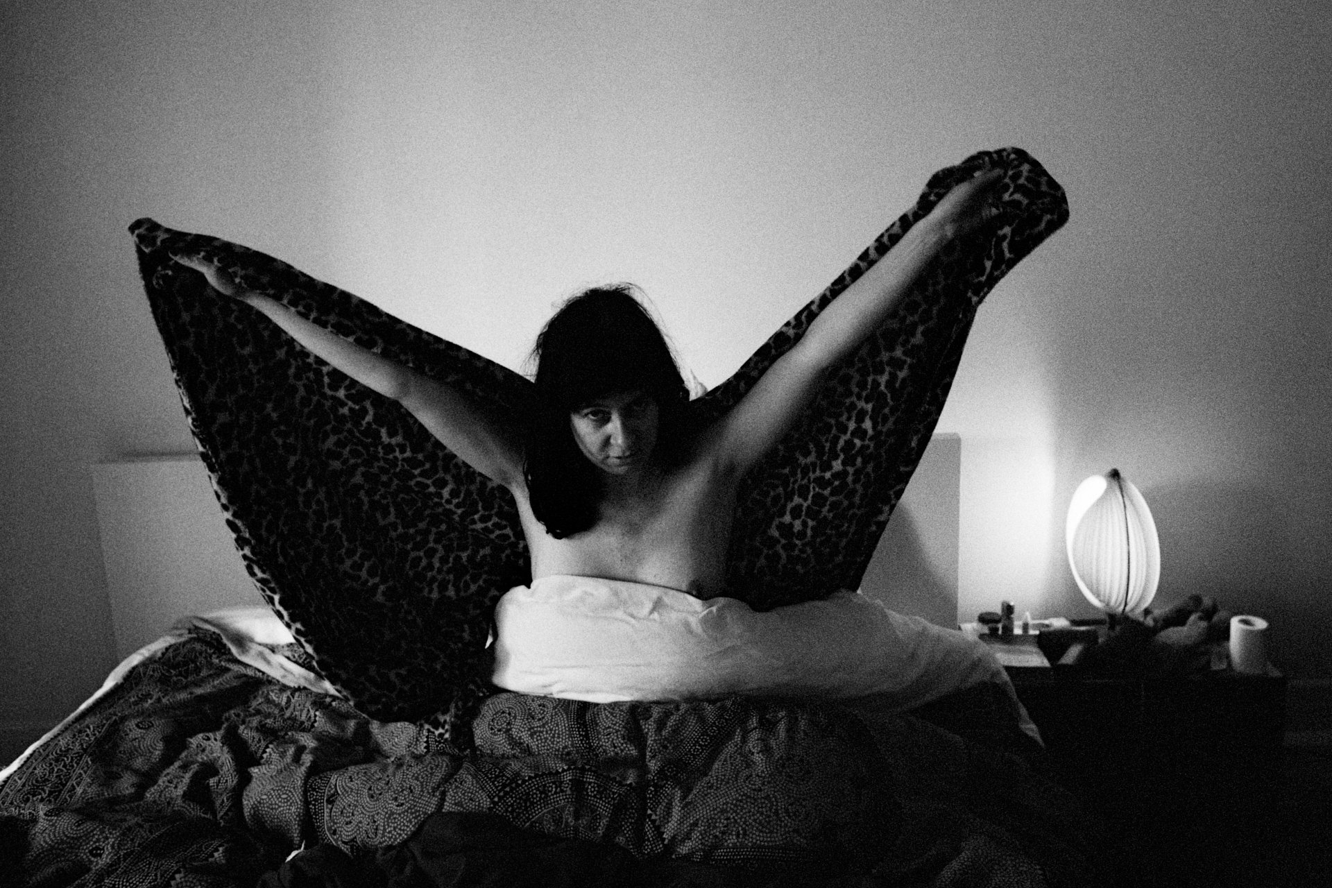 A woman sitting up in bed with her arms stretched out holding a leopard print fabric, looking down at the camera. The scene is in black and white, with a bedside lamp and clutter on the nightstand visible in the background.