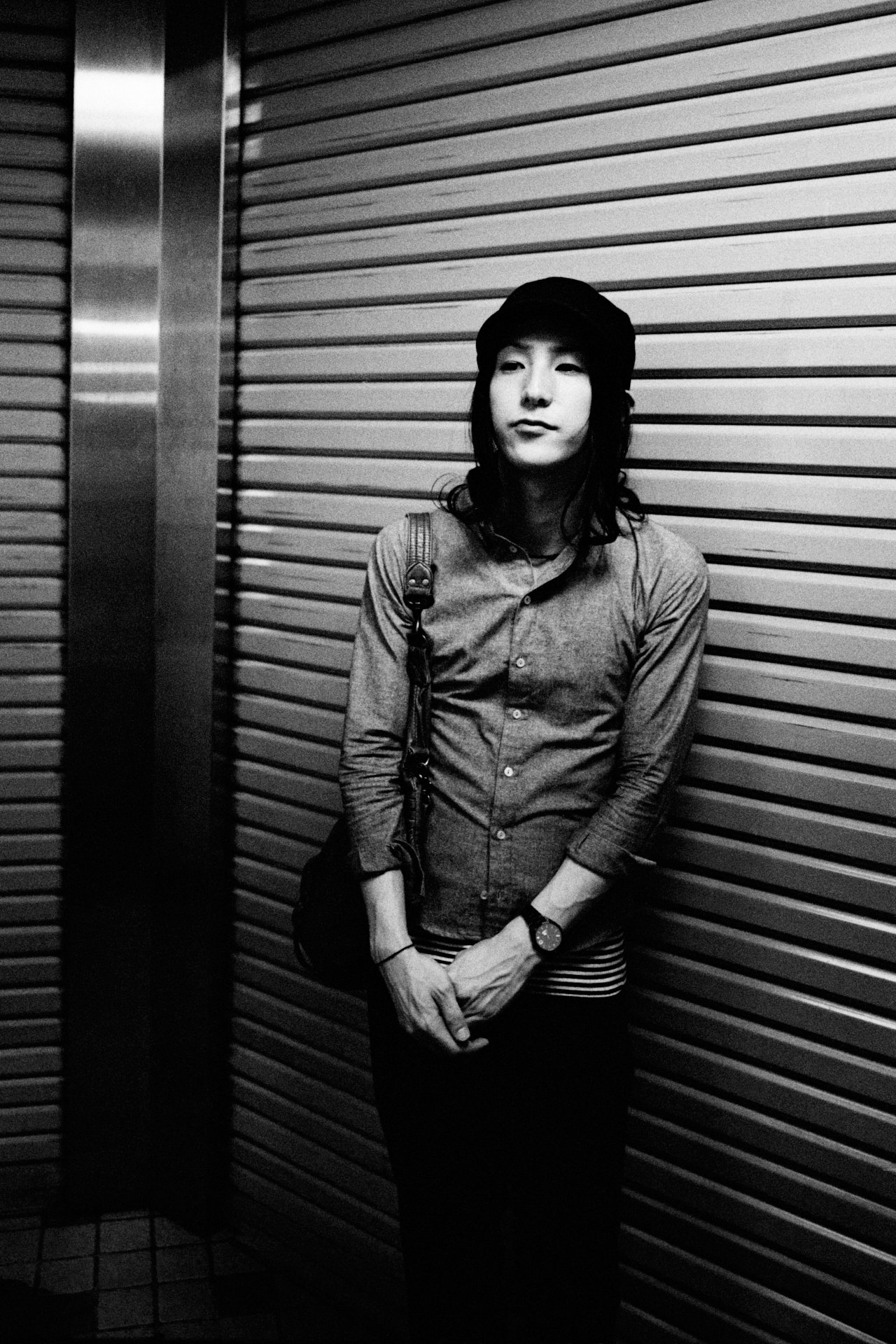 A woman standing against a metal, horizontal slat wall in an elevator or similar enclosed space, wearing a hat, long-sleeved shirt, and striped skirt, with a bag over her shoulder and a watch on her left wrist.