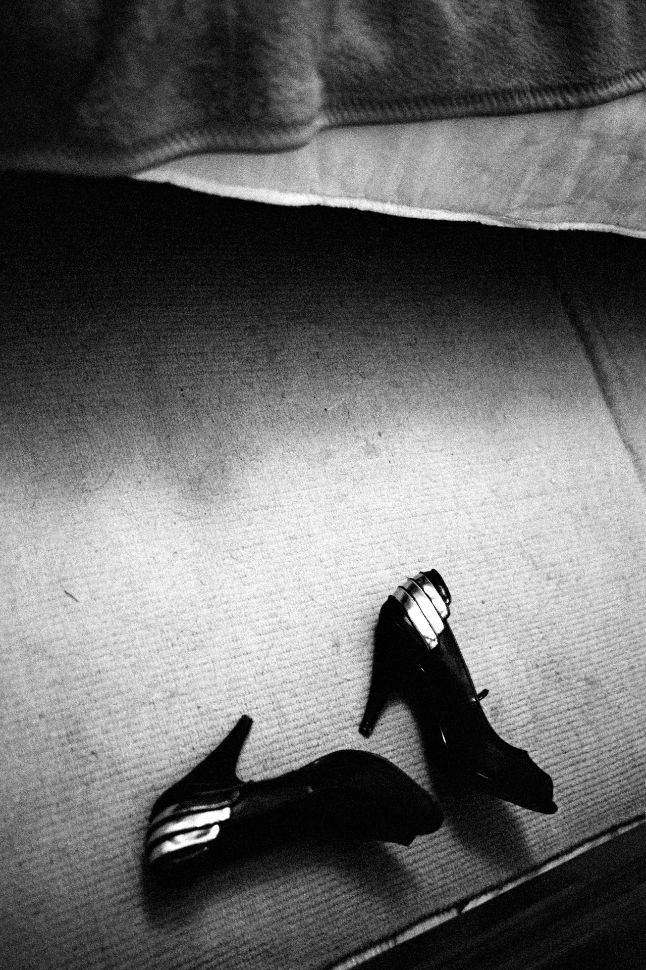 Black high-heeled shoes placed on a textured surface, with the background showing a bed with a blanket and bedsheet.