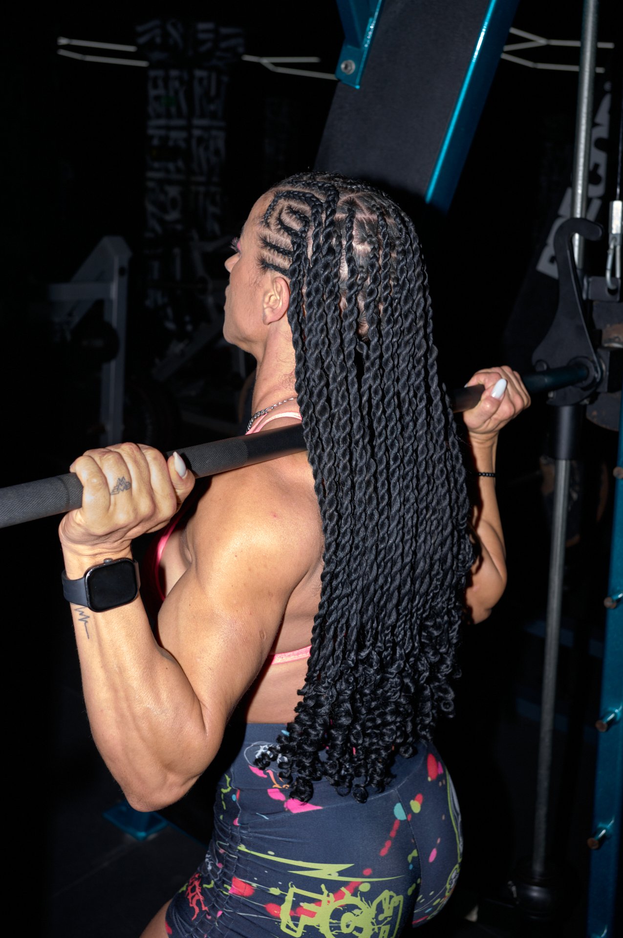 A woman with highlight-colored hair styled in long, twisted braids is lifting a barbell while working out in a gym.