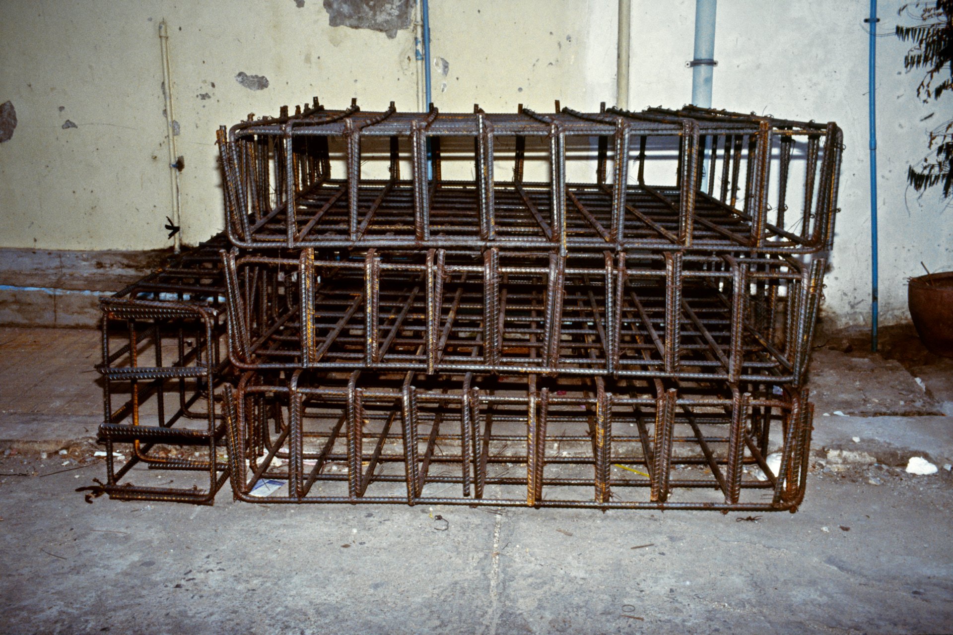 Stacked metal rebar cages in an unfinished building corner against a wall with peeling paint and exposed pipes.