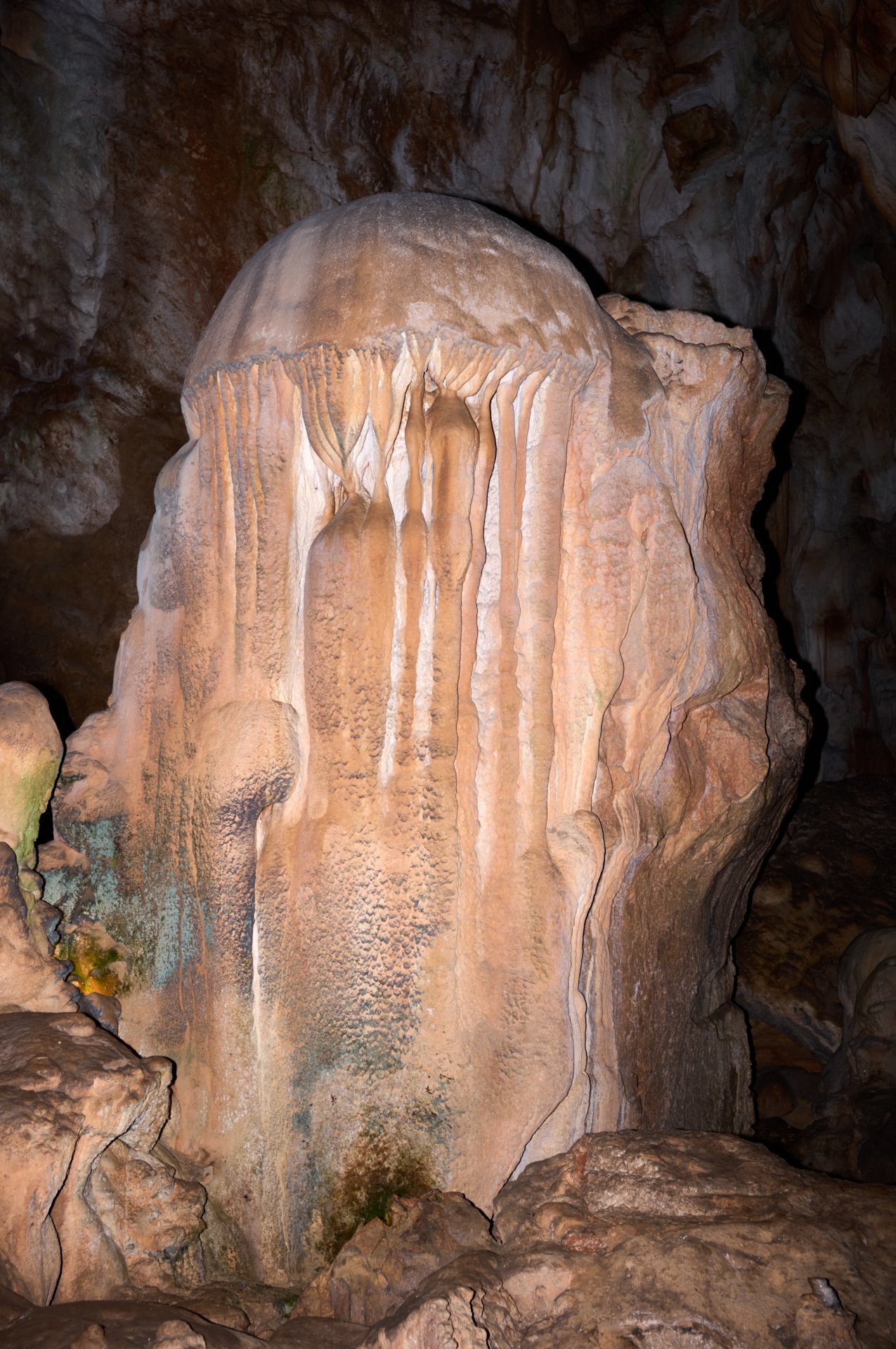 A large, colorful stalactite formation inside a cave, with dripping mineral deposits hanging from the ceiling and rugged rock surfaces surrounding it.