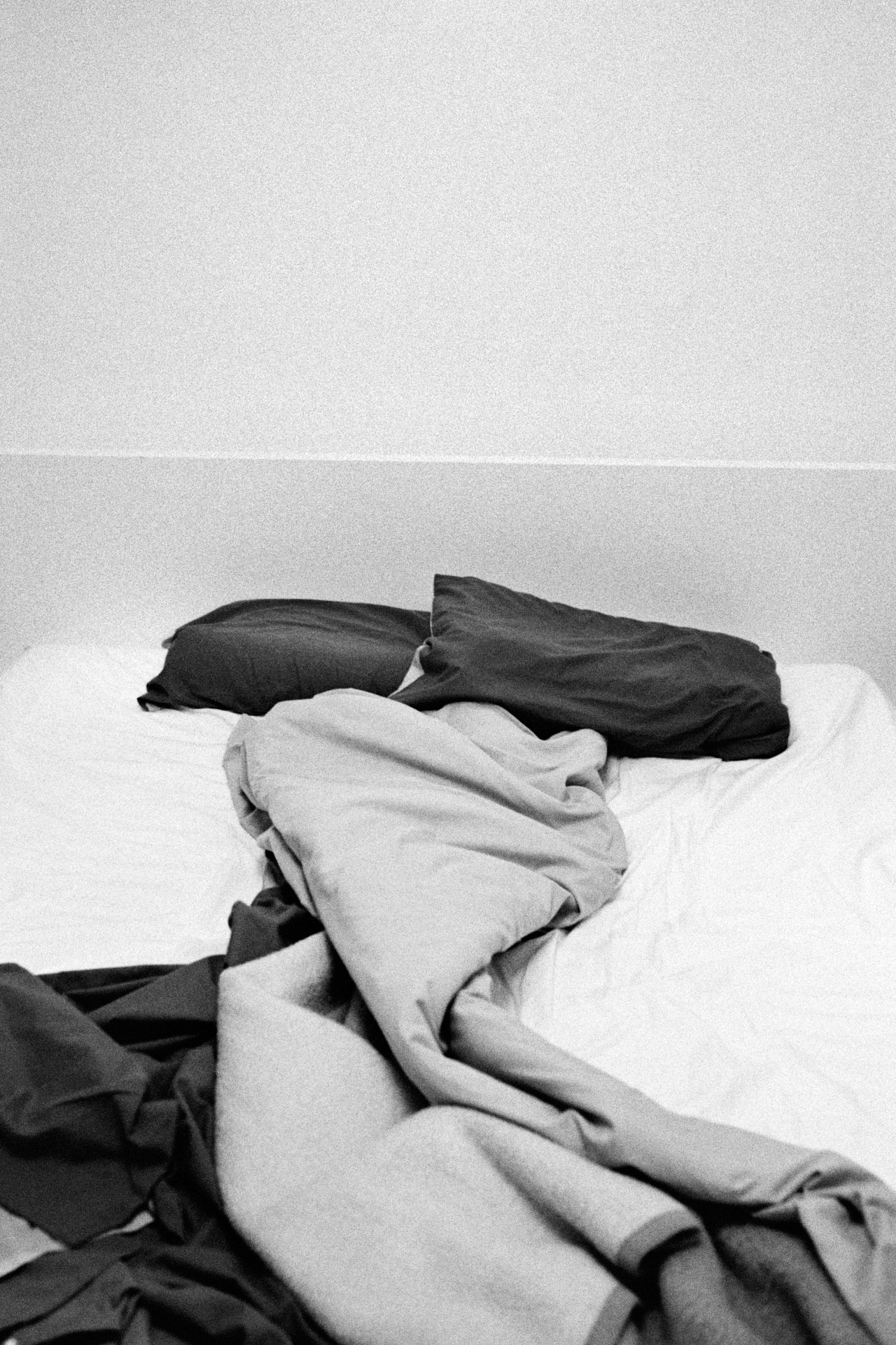 A messy bed with crumpled bed sheets, blankets, and pillows on a plain wall background.