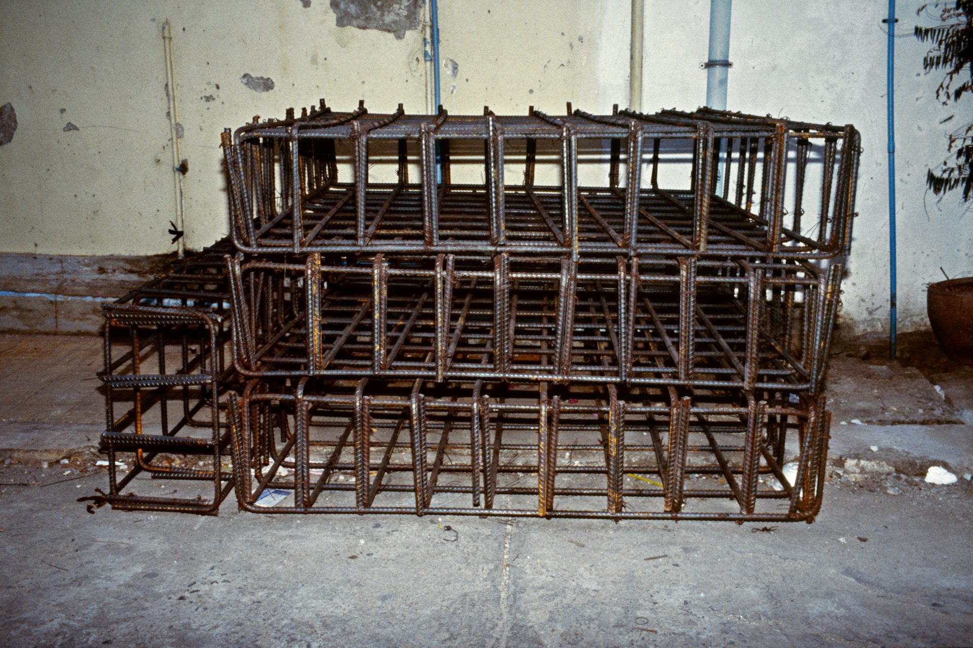 Stacked metal rebar cages for construction in an indoor space with a concrete floor and peeling yellow wall.