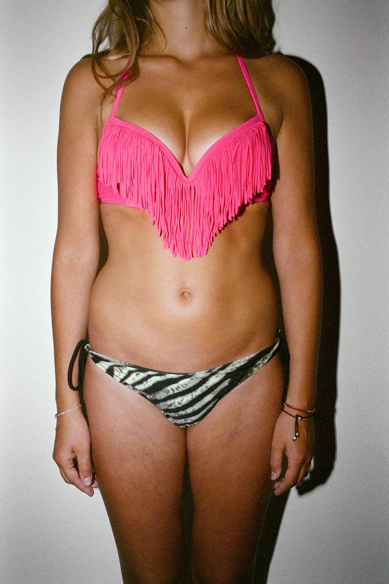 A woman wearing a pink fringed bikini top and zebra-print bikini bottoms standing against a light-colored wall.