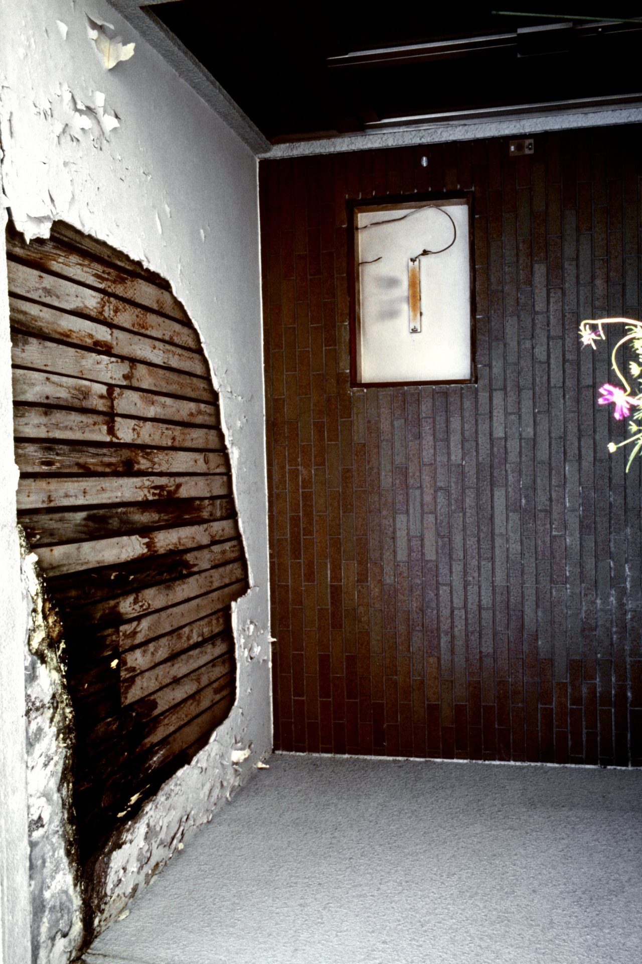 A room with damaged wooden paneling and peeling wallpaper on a white wall, with a brick wall in the background featuring an electrical panel with wires.