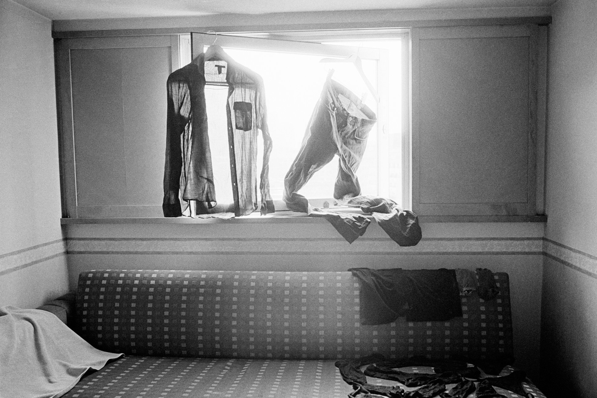 Clothes hanging on a window sill with sunlight coming through, and a bed with scattered clothes.