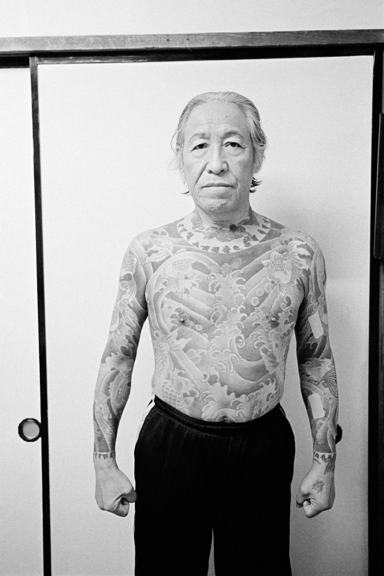 Black and white photo of a shirtless man with extensive tattoos on his chest, arms, and neck, standing in front of a plain white background.