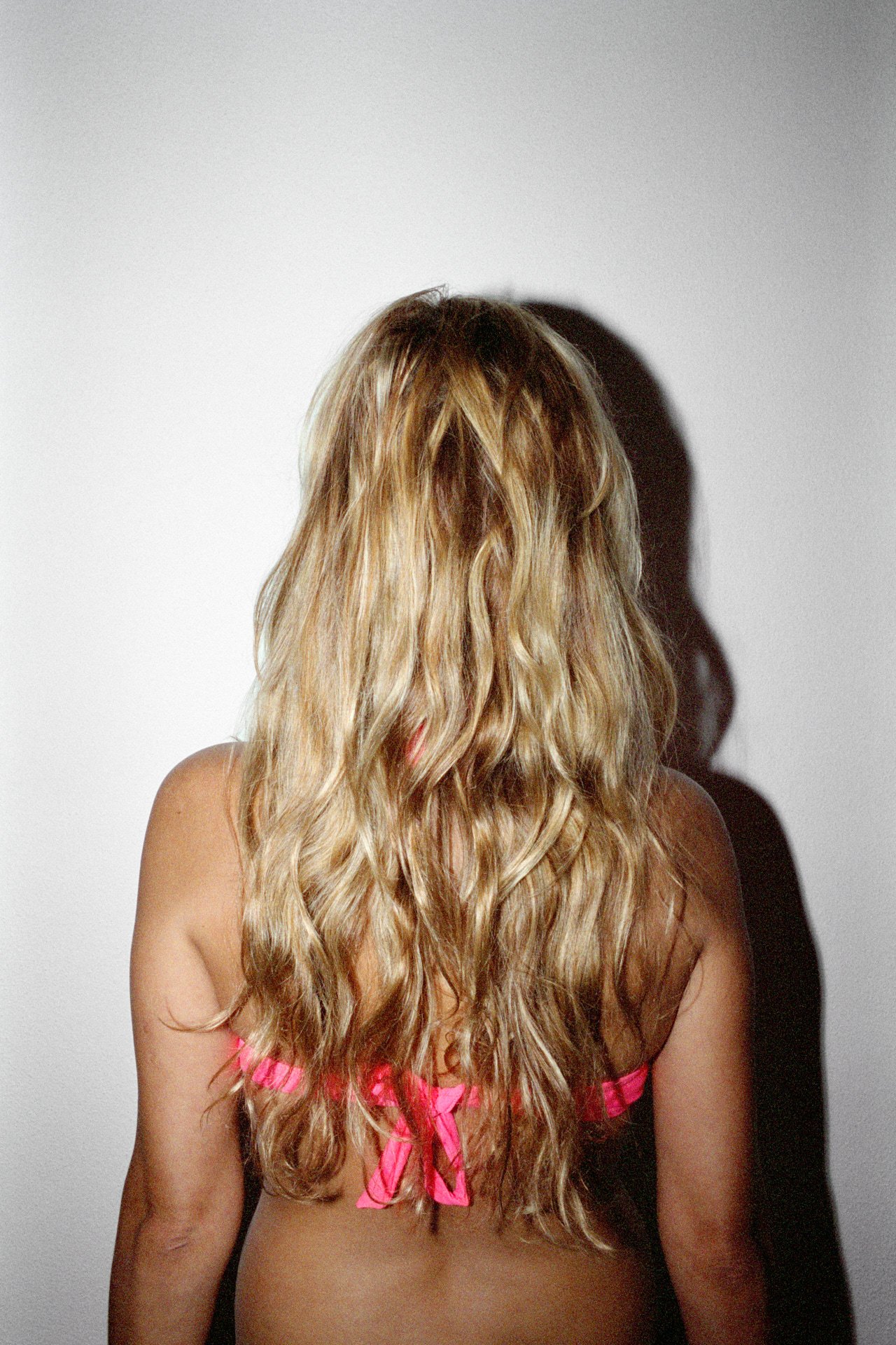 Back of a woman with long, wavy blonde hair, wearing a pink garment with pink ribbon bows, standing against a plain white wall, casting a shadow.