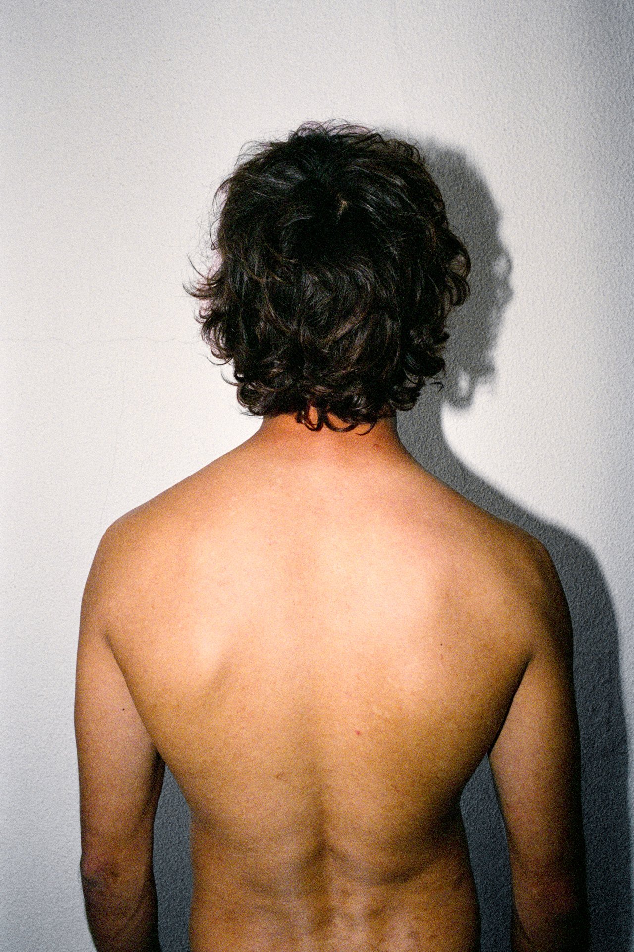 Back view of a person with dark curly hair and bare upper back standing against a plain white wall.