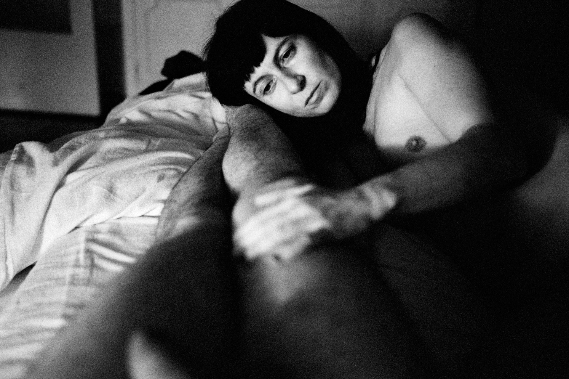 A black and white photo of a woman with dark hair and bangs lying on a bed, looking off to the side with a pensive expression.