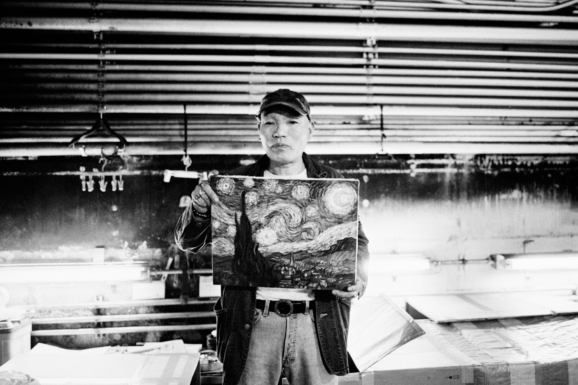 Black and white photo of a person wearing a cap and jacket, holding a painting of Vincent van Gogh's 'Starry Night' in a workshop or studio with ceiling beams and work tables.
