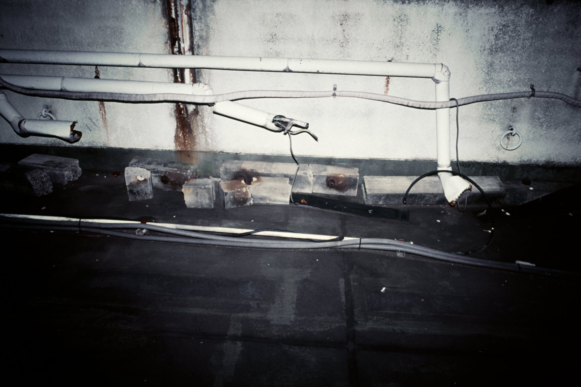A wall with peeling paint and rust stains, with multiple white pipes installed along the wall, some connected and some disconnected or damaged, with a black power cable hanging loosely.