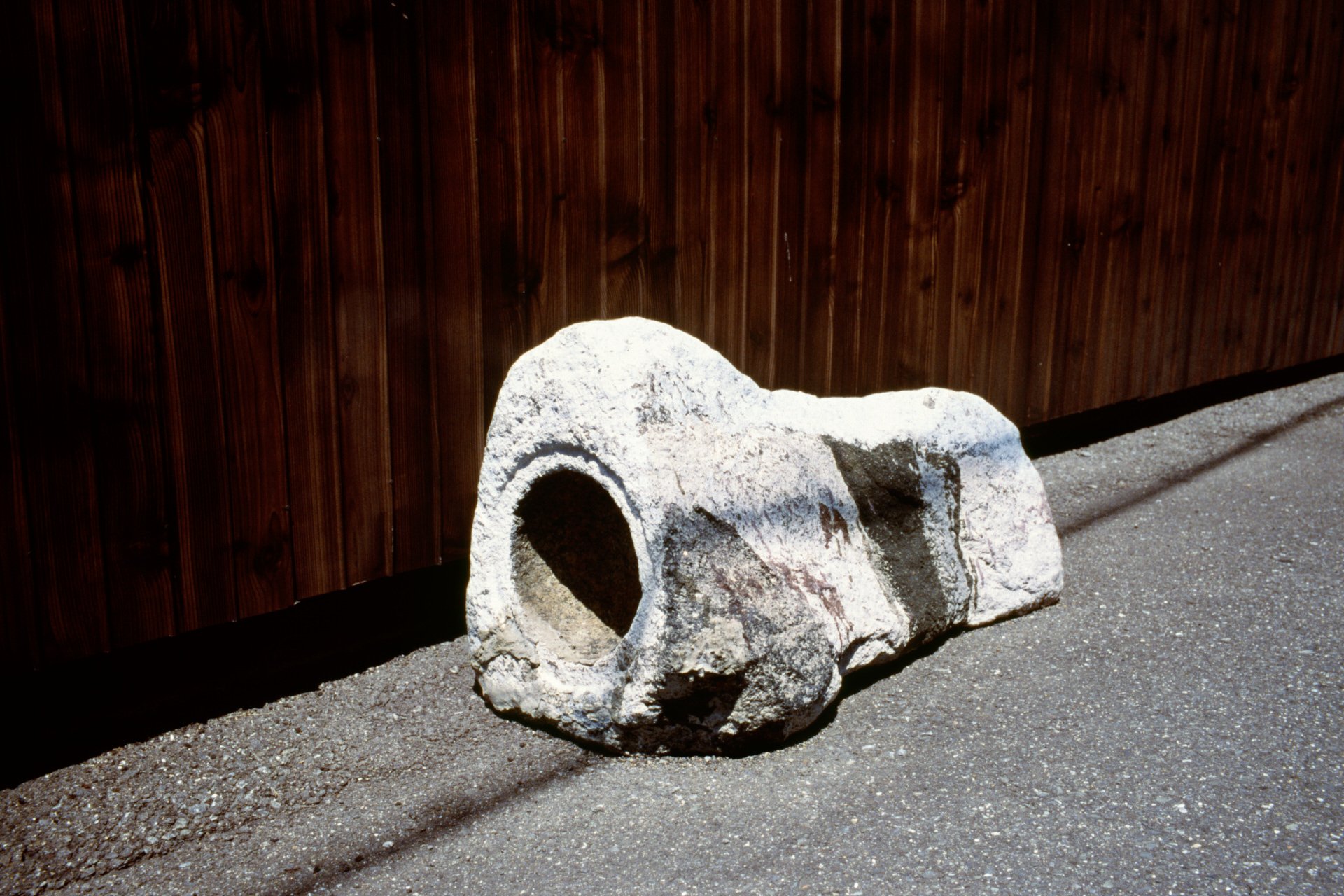 A large, weathered, hollow stone sculpture resembling a knee joint or bone, placed on a rough concrete ground near a wooden fence wall.
