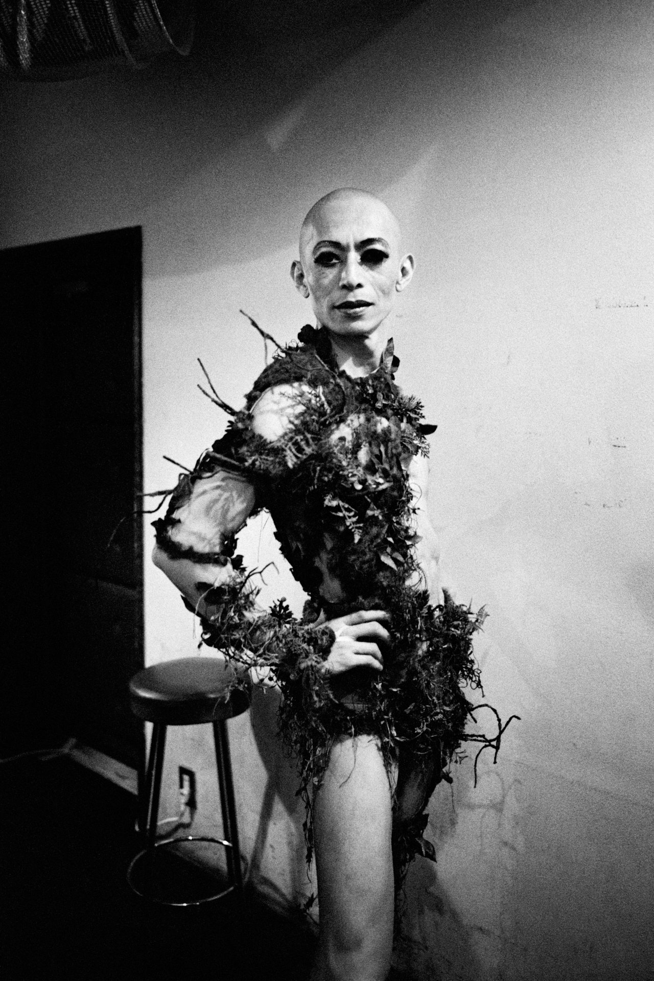 A bald woman with dark eye makeup wearing an outfit made of leaves and natural materials standing with one hand on her hip in a room with a stool, a chair, and a white wall.