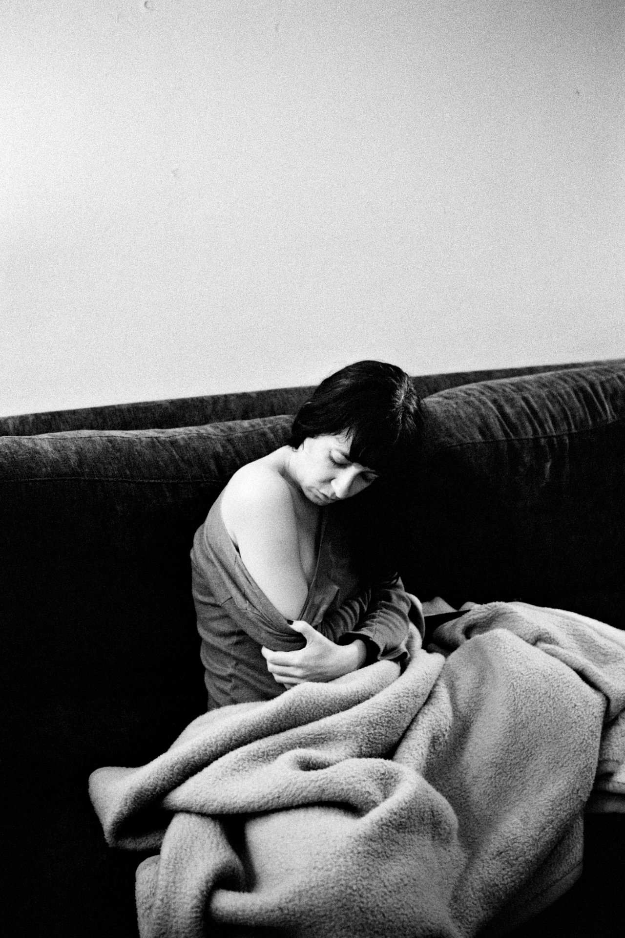 A woman sitting on a couch with a blanket, appearing sad or contemplative, in a minimalistic room with plain walls.