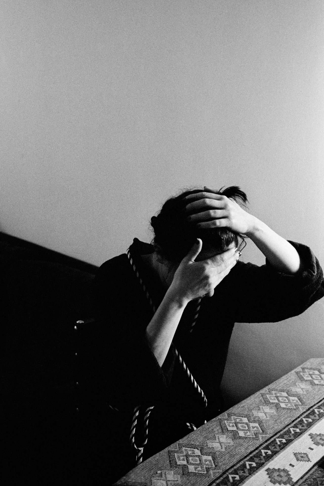 Black and white photo of a person with short hair and glasses, covering their face with their hands, sitting at a table with a patterned tablecloth.