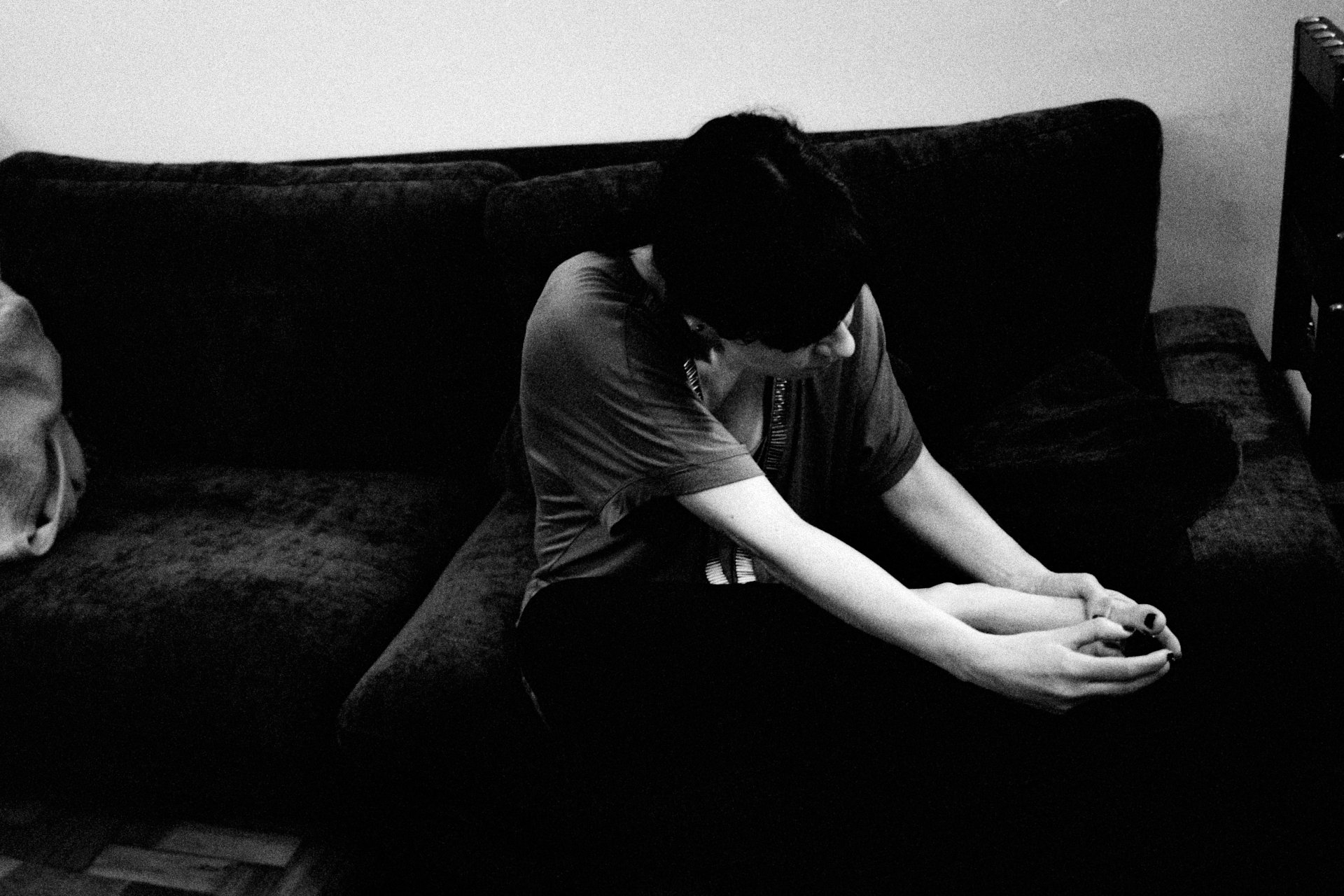 A person with short hair and glasses sitting on a couch, looking down at their hands, in a black-and-white photo.