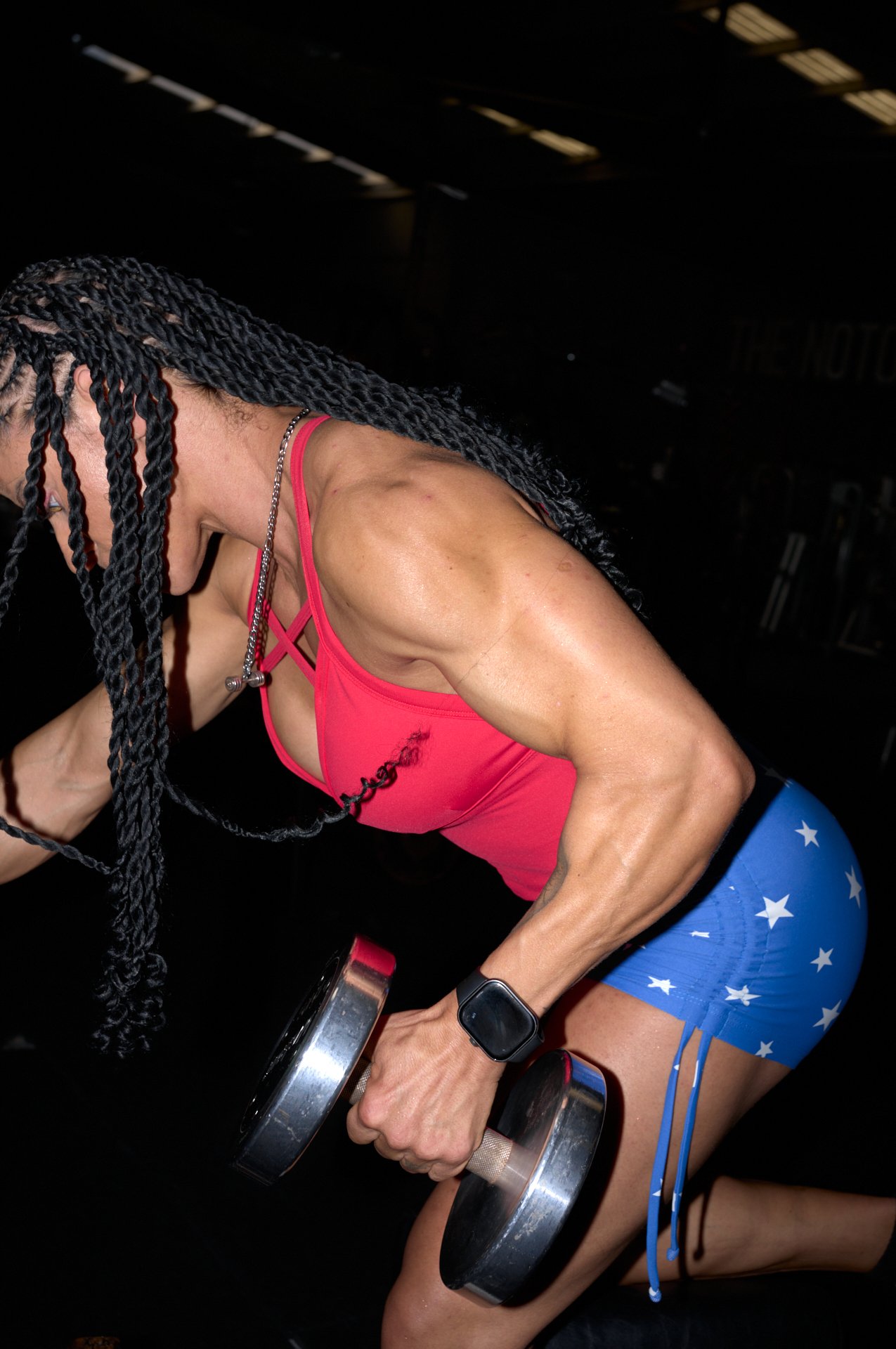 A woman with long braided hair works out in a gym, lifting a dumbbell in each hand while kneeling on the floor, wearing a pink sports top, blue star-patterned shorts, and a smartwatch.