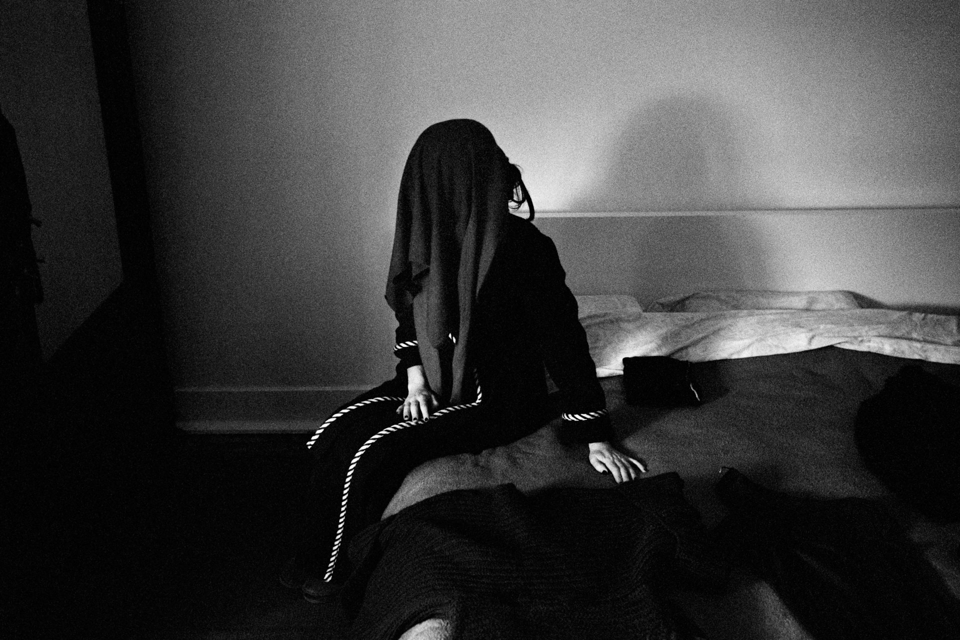 A person with long hair wearing dark clothing with white rope accents, sitting on a bed in a dimly lit room, facing away from the camera.