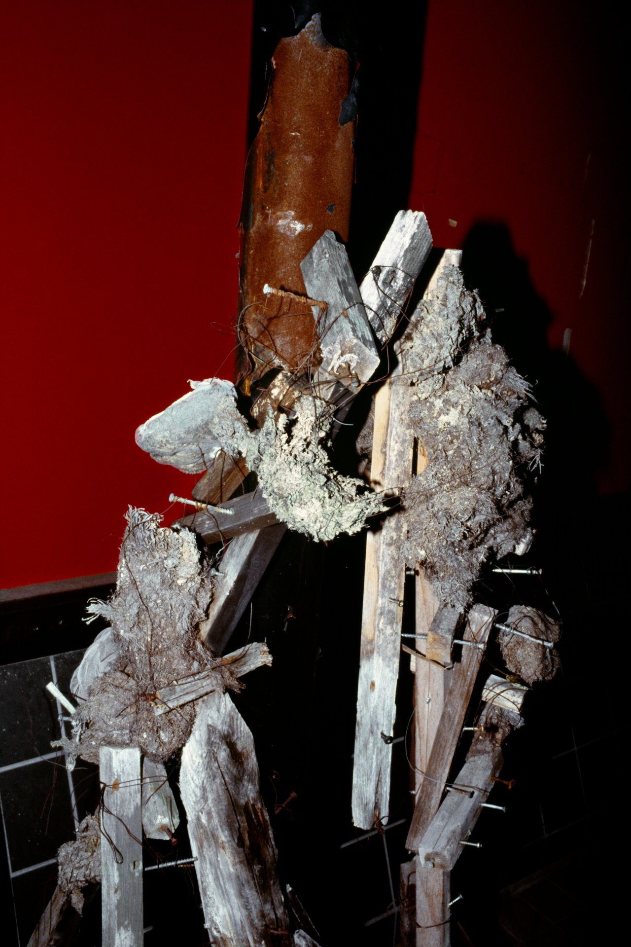 Close-up of broken wooden poles and debris tied together with wire, with peeling brown paint and a distressed appearance, against a dark red background.