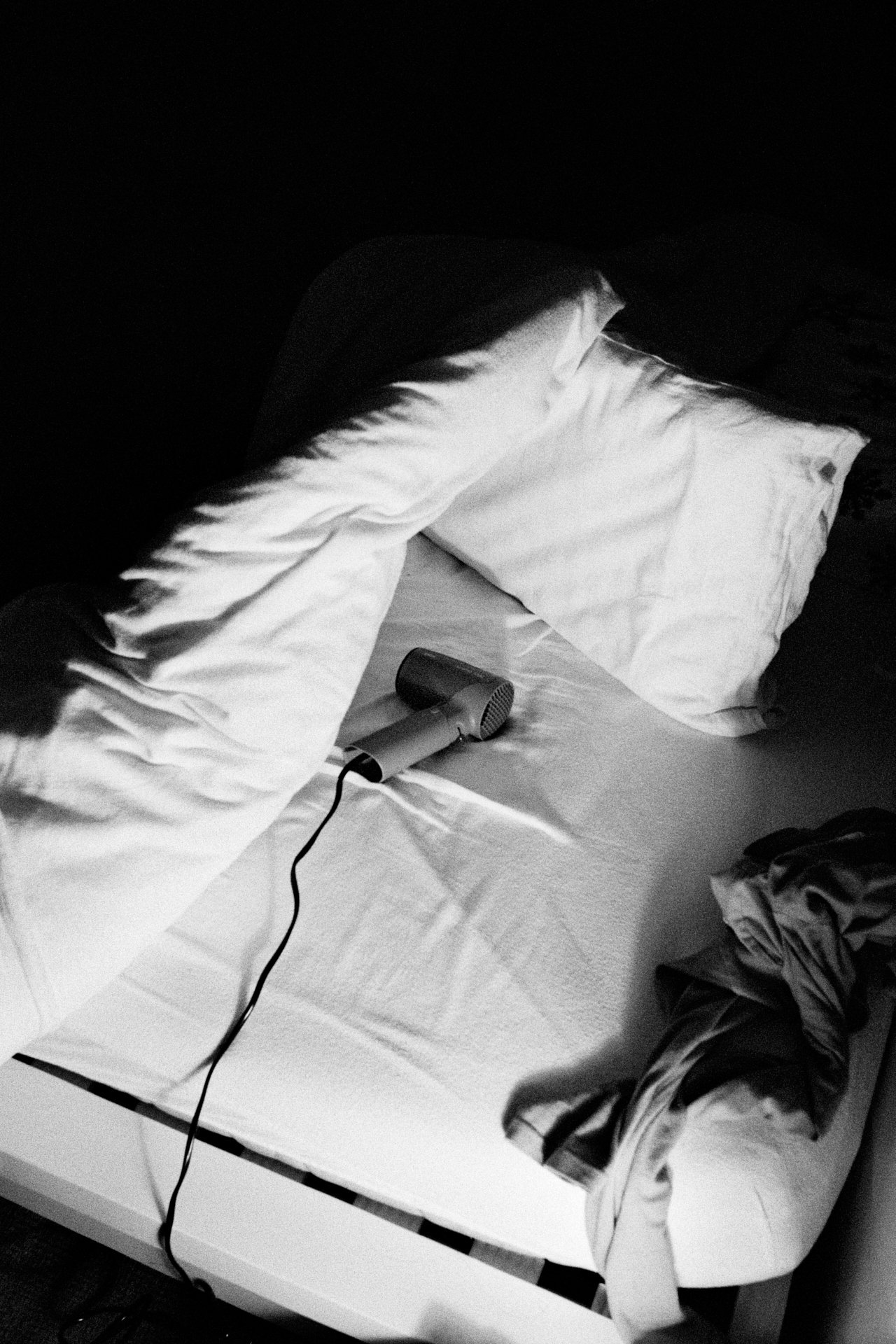 Unmade bed with a hair dryer on it, and clothing scattered nearby, in a black-and-white photo.