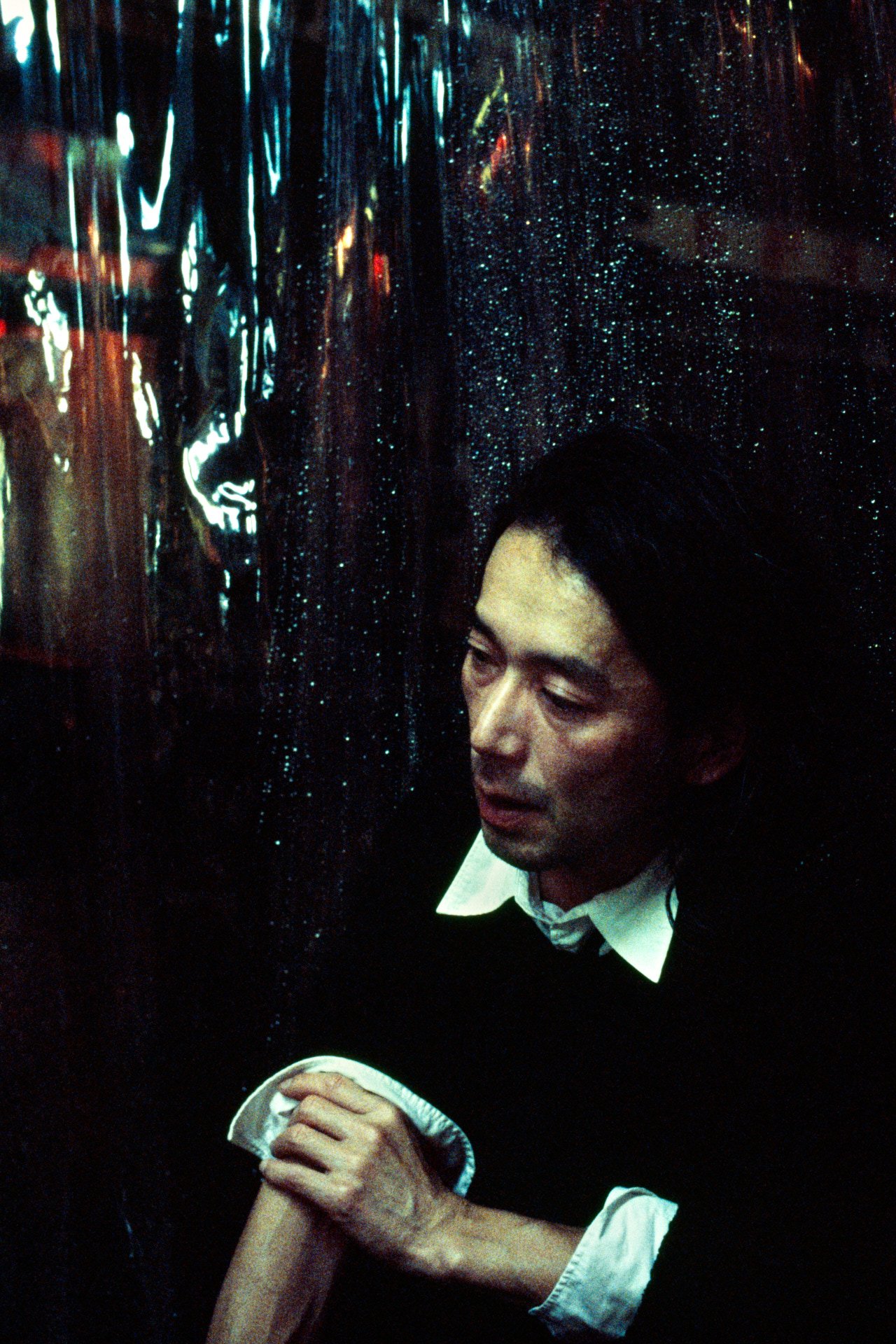 A man with short black hair, wearing a white shirt with a black jacket, appears to be praying or meditating with his hands clasped together against a dark, shiny, reflective background.