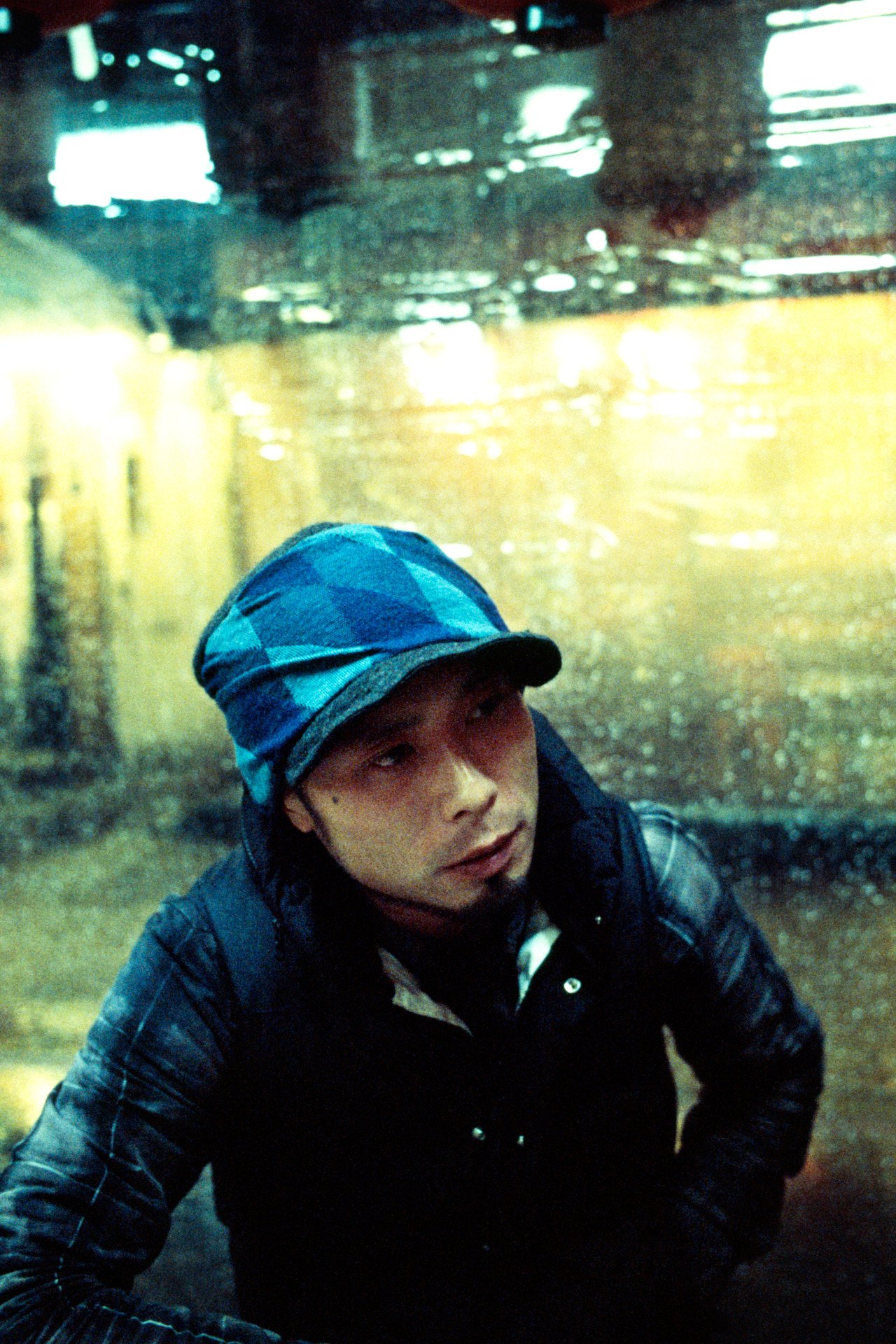 A person wearing a blue and black plaid cap and a black jacket, looking up in a rainy environment with a wet, dark background.
